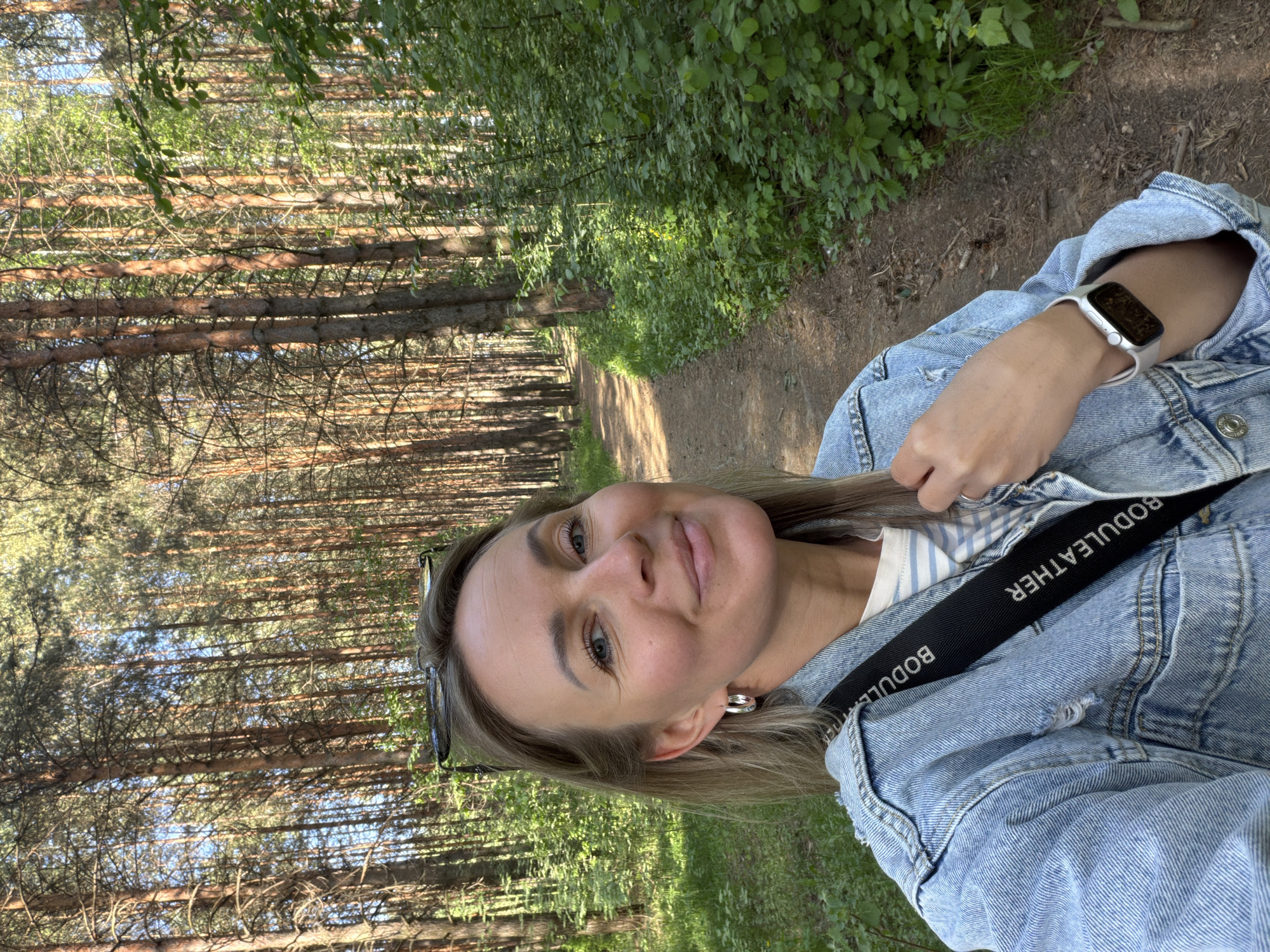 A woman stands on a path through a pine forest. She wears a denim jacket and a white Apple Watch on her left wrist. The strap of a BODULEATHER bag crosses her torso. Trees line the path on both sides, with sunlight filtering through the foliage. The scene is likely in a forest area within or near the city of Chaikovsky, Russia, although there is no specific identifier of that city in the image.