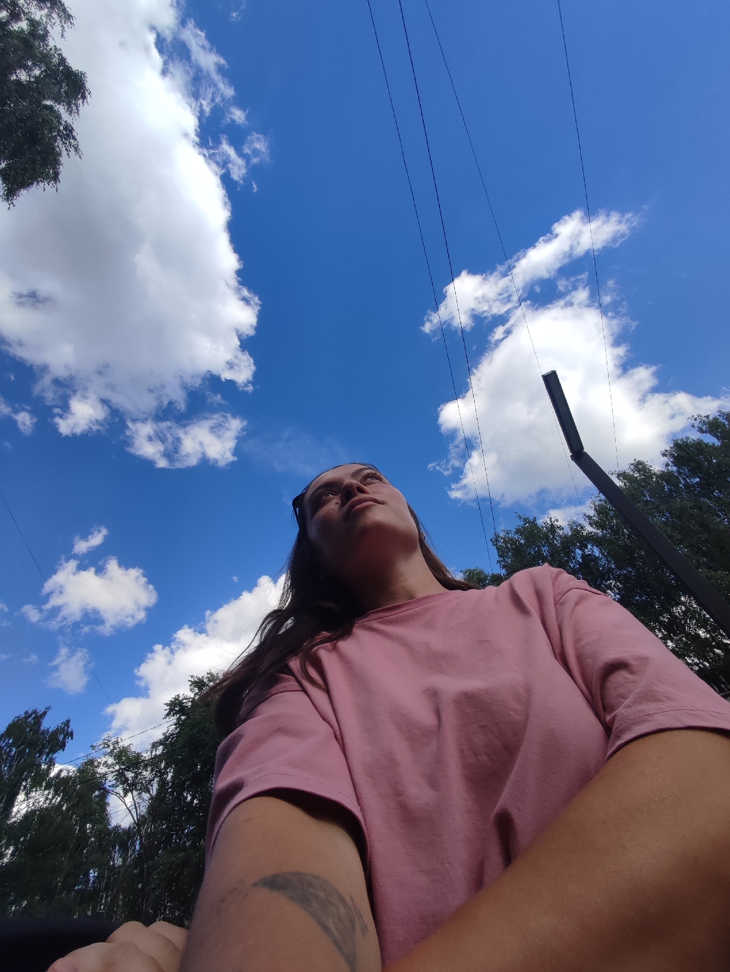A low-angle shot depicts a young woman looking up at a bright blue sky dotted with fluffy white clouds. She is wearing a pink t-shirt and has her hair pulled back. The image shows power lines stretching across the sky, and the tops of trees and a utility pole are visible in the background. The scene appears to be outdoors, likely in a park or open area within the city of Chaikovsky, Russia, given the presence of trees and infrastructure.