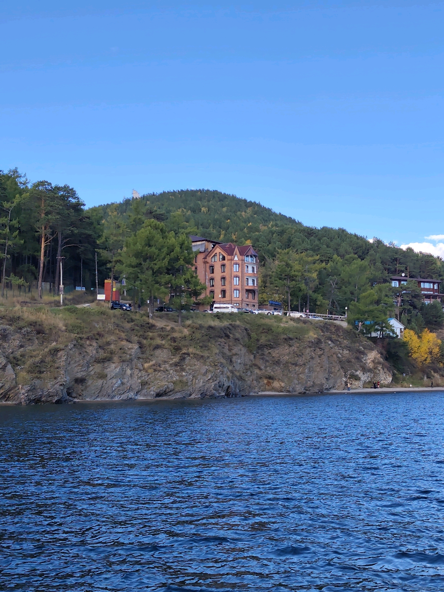 This image depicts a scenic view of Lake Baikal in Listvyanka, Russia. In the foreground, the deep blue water of the lake is visible, with gentle ripples on its surface. A rocky cliff face rises from the water's edge, covered in sparse vegetation and some trees. On top of the cliff, a multi-story brick building with a reddish-brown facade and a dark roof stands prominently. This building has a modern design with numerous windows. Adjacent to it, a dense forest of evergreen trees covers the hillside, with a few deciduous trees displaying yellow foliage, indicating autumn. Several vehicles are parked along a road or pathway on the cliff. A small white building with a dark roof is situated further down the hillside. In the distance, more buildings are nestled among the trees, and the sky is a clear, bright blue. A few people are visible on a small beach area at the base of the cliff. The overall scene is one of natural beauty combined with human habitation, suggesting a resort or residential area.