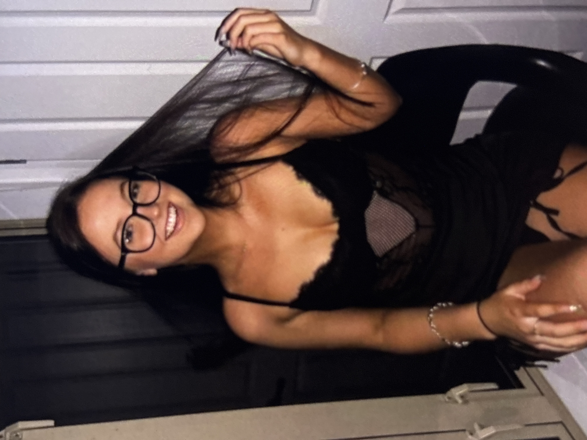 A young woman with long, dark hair and glasses is sitting in a chair. She is wearing a black lace outfit. Her right hand is holding her hair and her left hand is resting on her leg. The background is dark with some white doors on the right side.