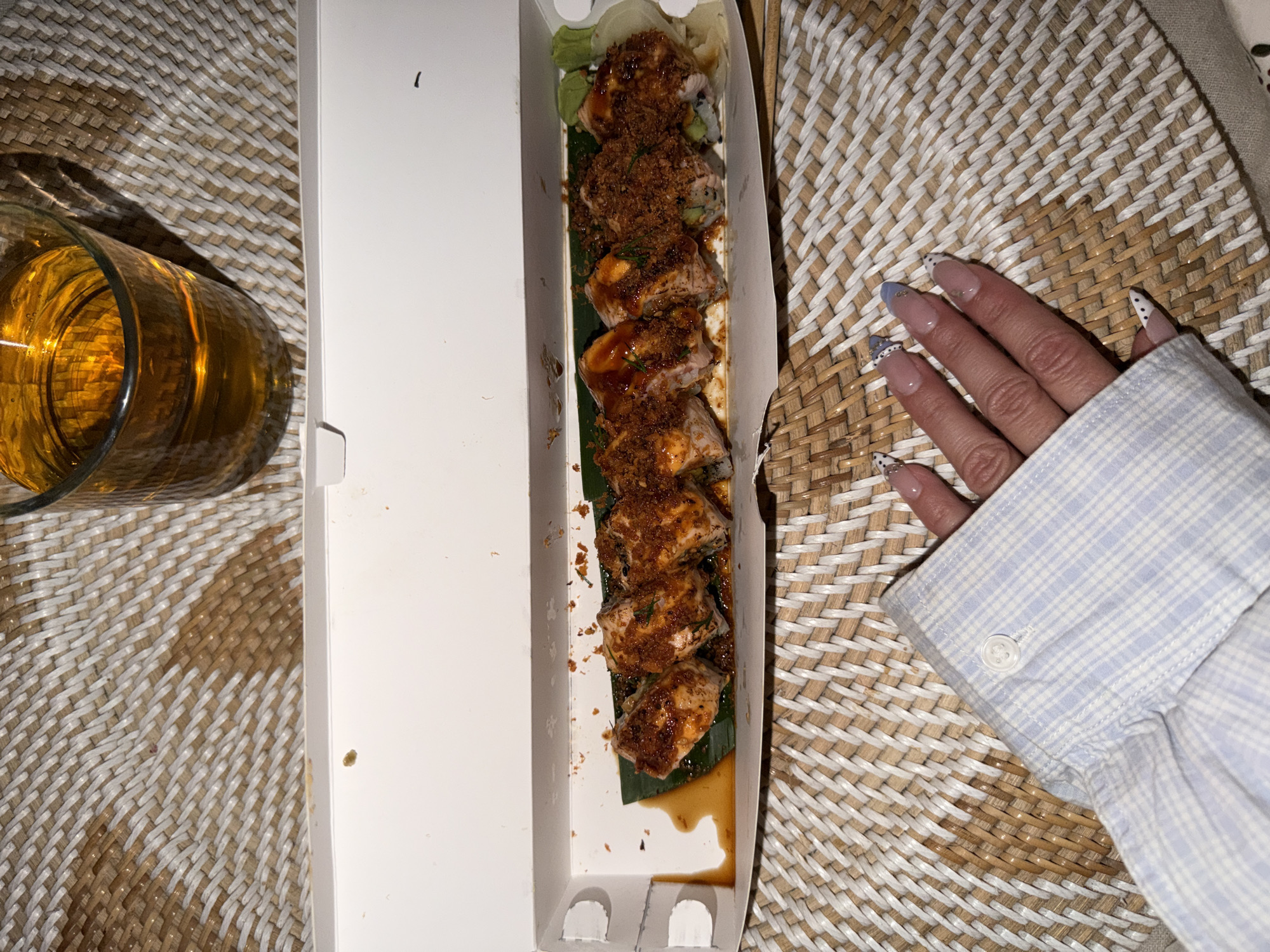 The image shows a top-down view of a partially opened white takeout container filled with sushi rolls. The sushi is topped with a dark sauce and sprinkled with brown bits. A glass of amber-colored liquid is visible to the left of the container. A hand with decorated nails rests on the right side of the image, near the edge of the container. The background is a woven surface of alternating light and dark colors. The photograph was taken in North Kuta, Indonesia.