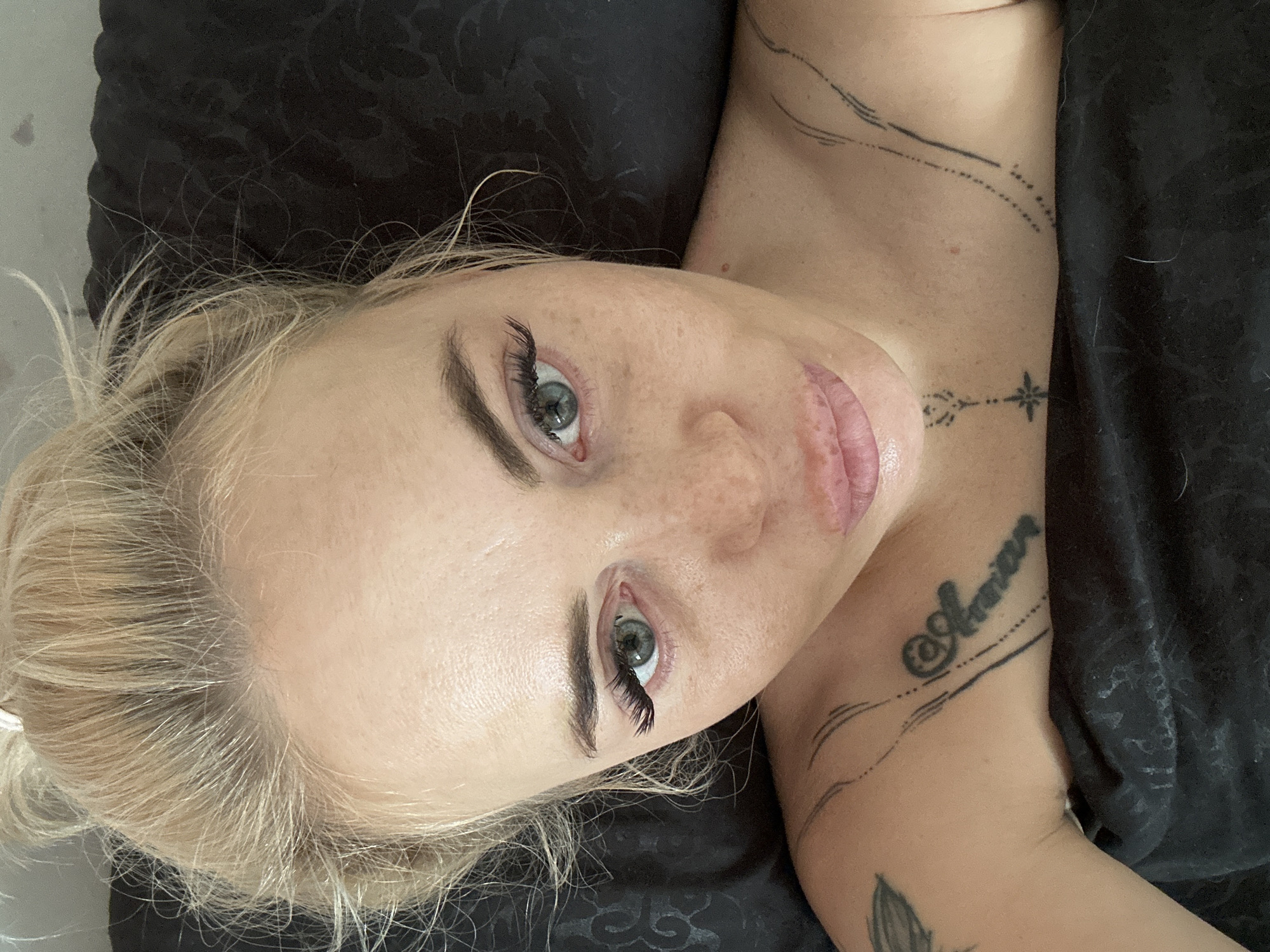 A close-up, overhead photograph captures an adult female lying down in an indoor setting, likely a bed, in the city of Unknown, Unknown. She looks directly into the camera with light-colored (grey-green) eyes, adorned with prominent false eyelashes. Her blonde hair is pulled back into a messy bun, with loose strands framing her face. Her complexion is fair with numerous visible freckles across her nose and cheeks. Her lips are full and slightly parted. Visible tattoos include a stylized "@Amor" on her left clavicle, a small geometric star-like design with a dotted line on her sternum, and multiple curved lines on her right clavicle. She is partially covered by a black fabric, possibly bedding, which has a subtle floral or damask pattern visible on the right side.