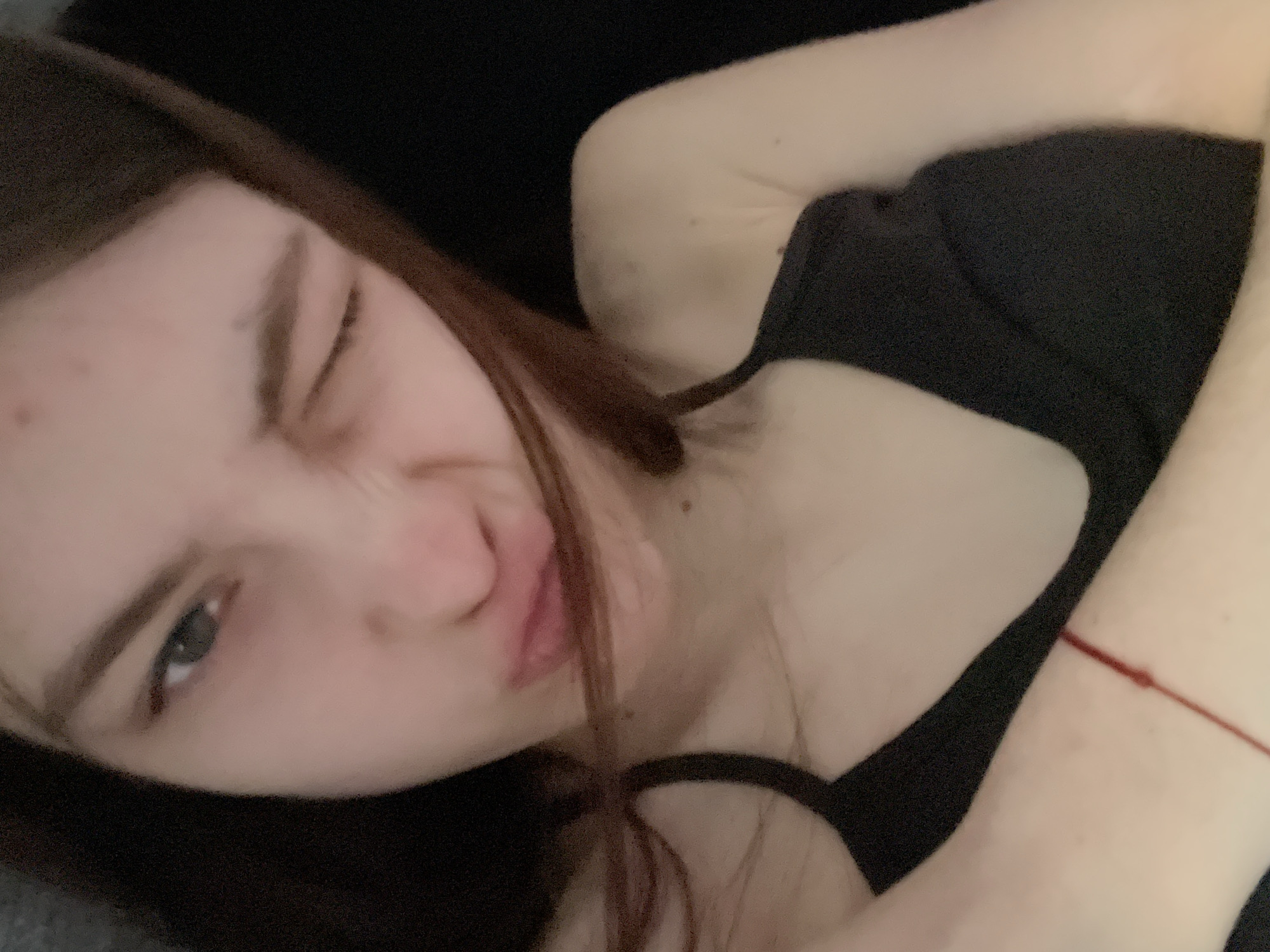 A young woman with long brown hair is lying down, wearing a black top with a cutout. She is winking one eye and puckering her lips. The background is dark, possibly a couch or bedding. A red string bracelet is visible on her wrist. The location is not discernible from the image.