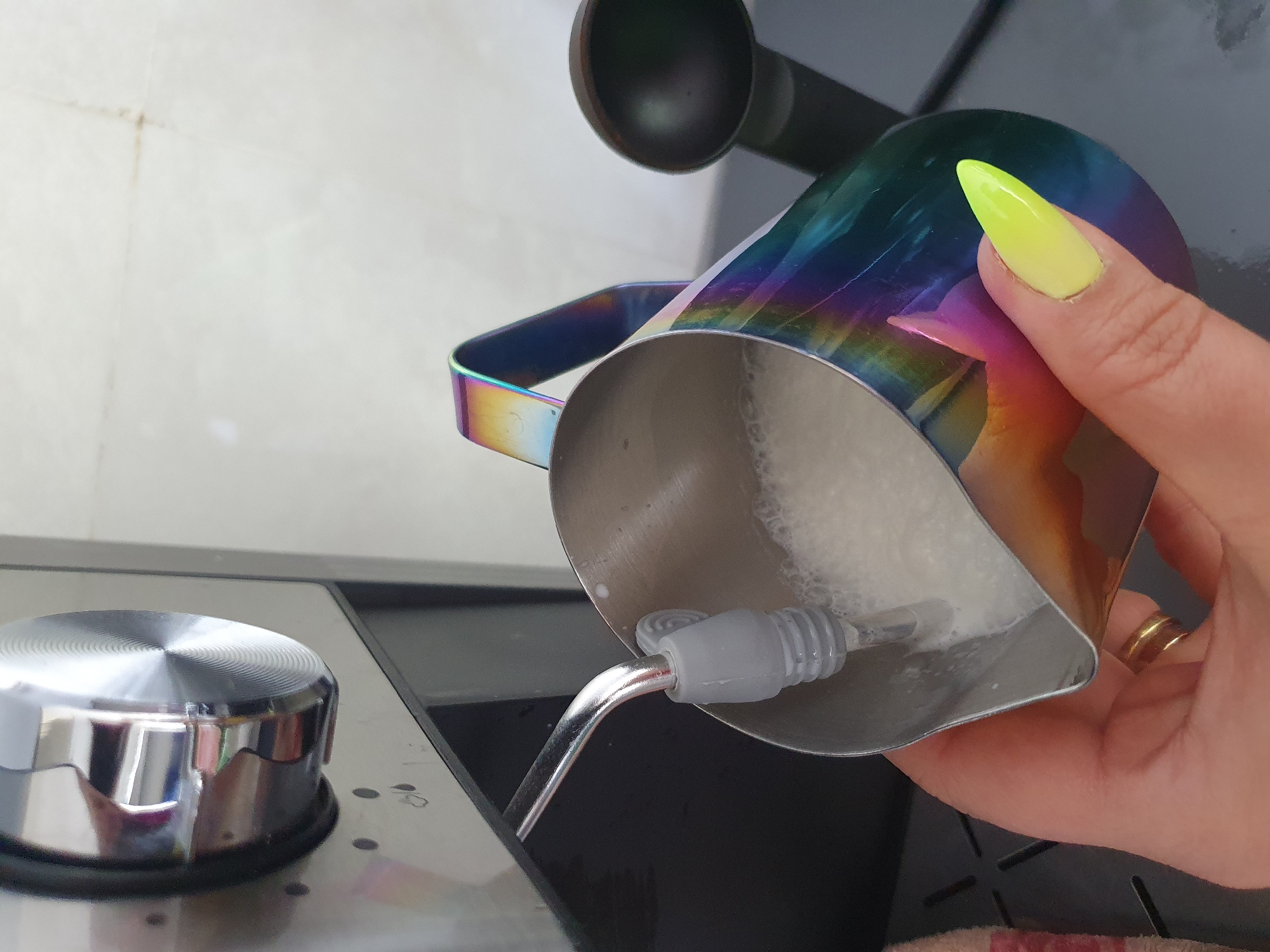 In Kheyrabad, Iran, an espresso machine's steam wand is frothing milk in a rainbow-colored stainless steel pitcher held by a woman with a neon yellow stiletto nail. The scene is indoors, likely a kitchen, with a tiled wall in the background. The pitcher is partially filled with frothed milk, and a small amount of coffee is being poured into it.