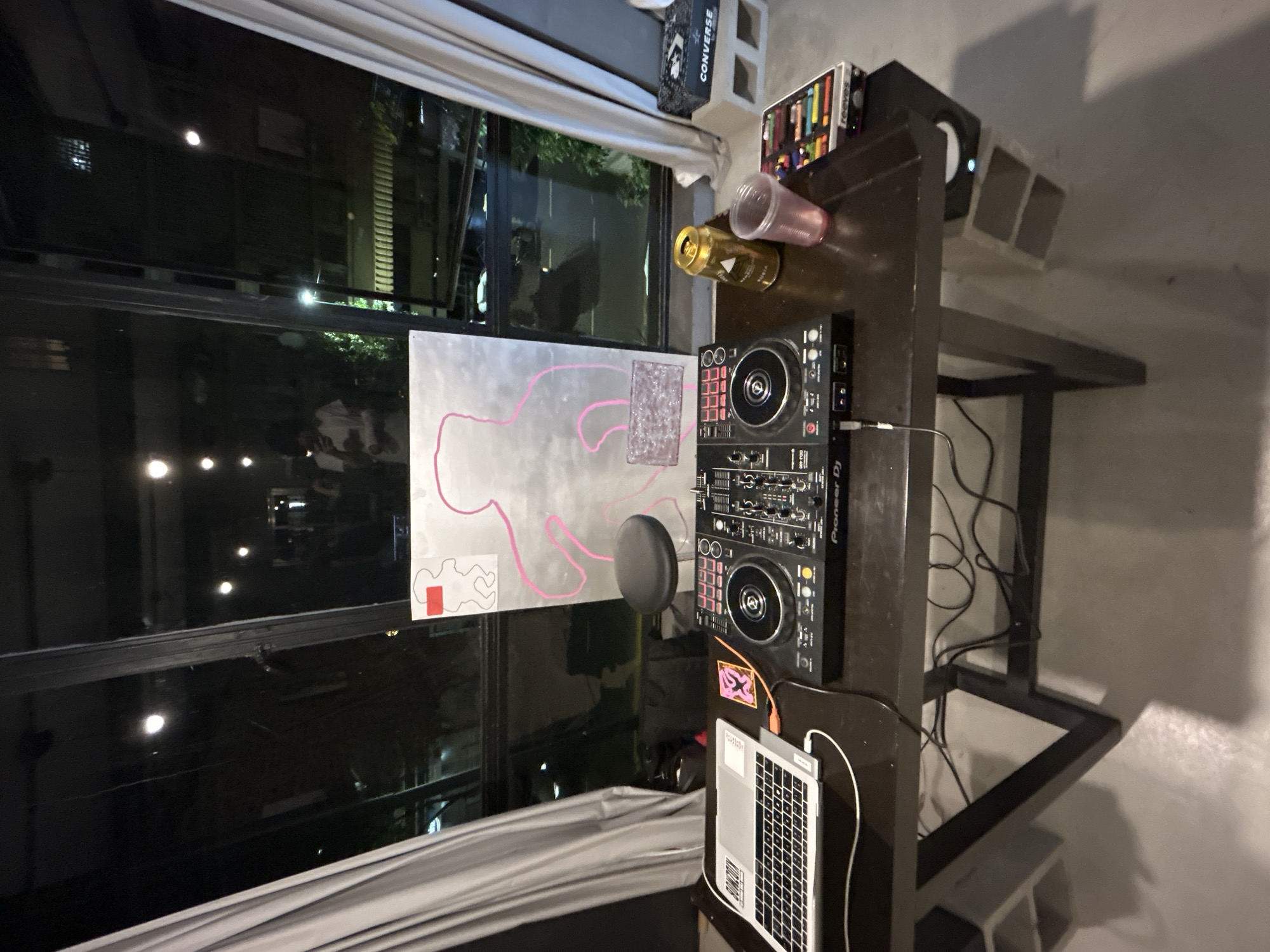 A high-angle, indoor shot shows a DJ setup and a window reflecting a night scene. The room appears to be in Buenos Aires, Argentina, indicated by a poster showing an outline of the country.
The DJ setup is centered on a dark-colored table or stand. It includes a Pioneer DJ controller, a laptop, and a microphone. Nearby are drinks (a can and a cup), a case of art supplies, and speakers resting on concrete blocks.
Behind the table is a large window covered by a curtain, providing a view of the city at night. The window reflects the interior of the room, including a person, adding depth to the image.