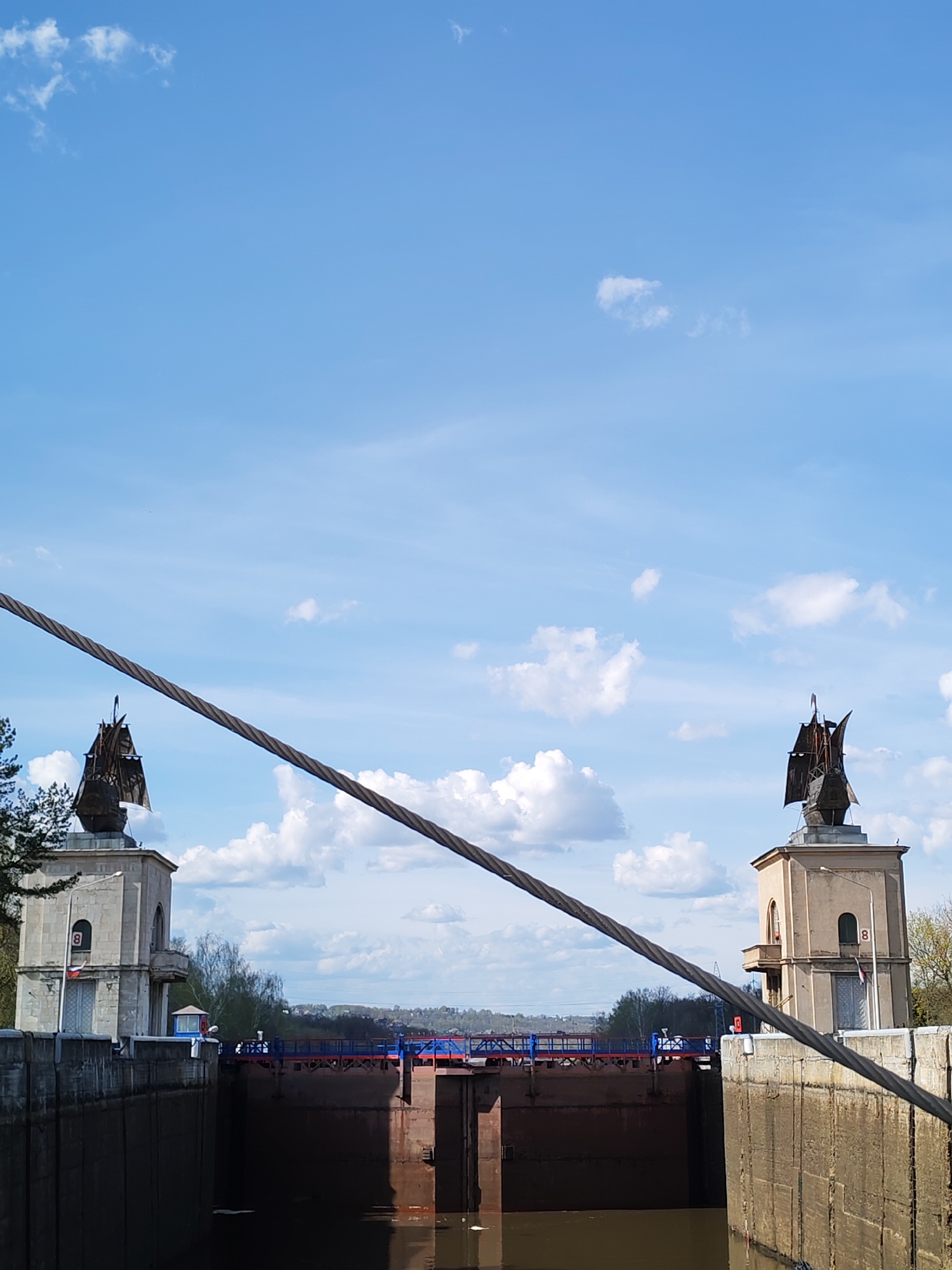 The image shows a view of the lock #8 on the Moscow Canal in Yakhroma, Russia. Two towers with ship sculptures on top flank the lock chamber. A thick cable runs diagonally across the frame. The lock itself is filled with dark water, and a blue and red pedestrian bridge spans the top of the chamber. Beyond the lock, a landscape with trees and buildings is visible. The sky is blue with scattered clouds.