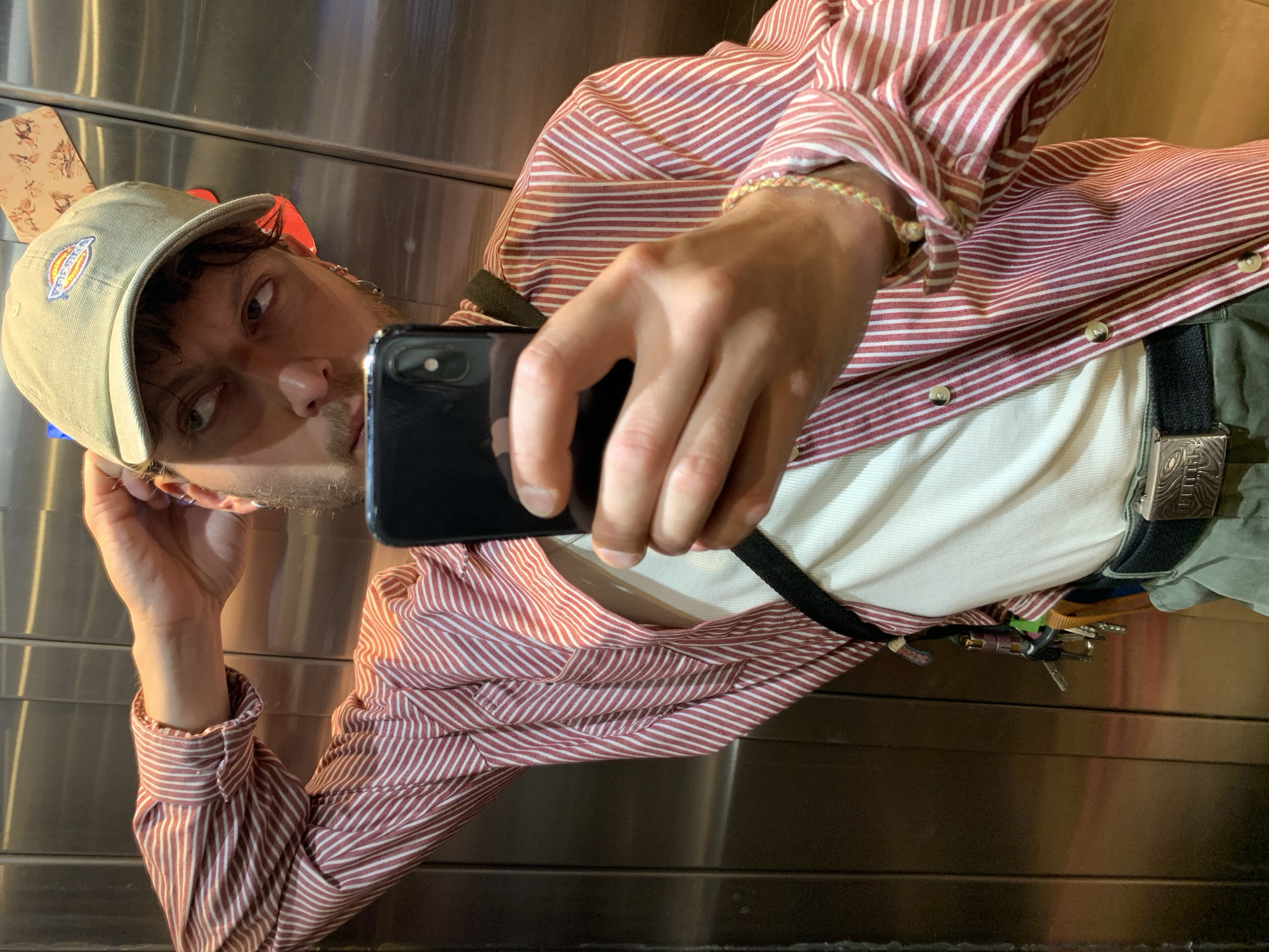 A man takes a selfie in an elevator with metallic walls. He is wearing a Dickies baseball cap, a red and white striped shirt over a white t-shirt, and olive green pants with a patterned belt buckle. Keys hang from his belt. The background suggests he might be in an elevator. The location in Tel Aviv, Israel cannot be determined from this image.