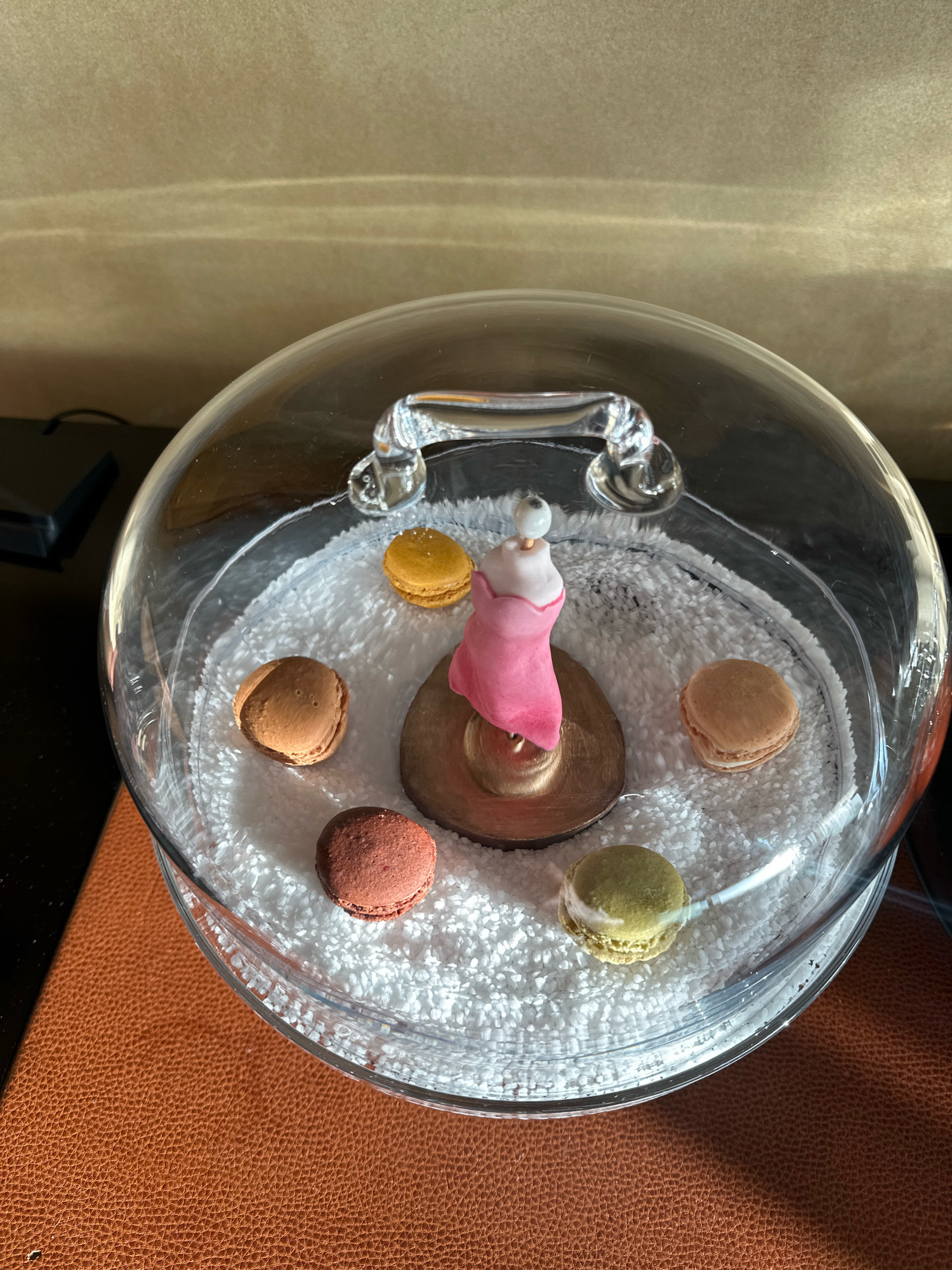 A clear glass dome covers a decorative arrangement on a textured brown surface. Inside the dome, white granular material forms a base for six macarons, each a different color: yellow, light brown, dark brown, red, tan, and green. In the center of the macarons, a small, pink, doll-like figure with a white head stands on a metallic base. The dome has a clear handle at the top. The background is a blurred, light-colored wall. This scene is not identifiable as being in London, United Kingdom.