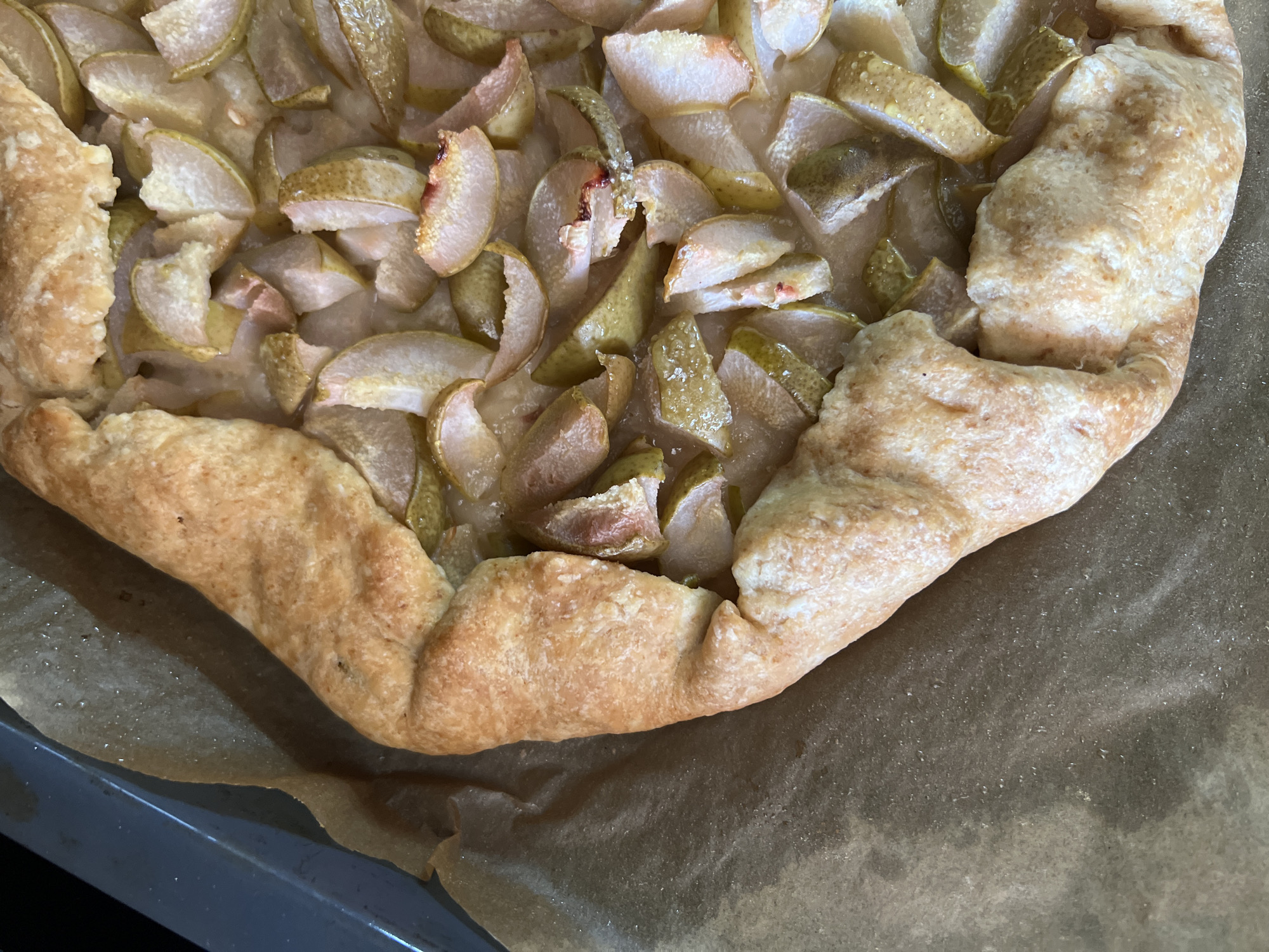 A close-up overhead shot captures a freshly baked pear galette resting on a baking sheet lined with parchment paper. The galette features a rustic, golden-brown crust folded over a filling of thinly sliced pears, glistening with syrup. The pears are a light green and yellow, with some showing reddish-pink edges from the skin. The crust is irregular and textured, with some parts appearing slightly darker and crisped. The background shows a dark grey metal baking tray on the left and the texture of the parchment paper on the right. The scene is brightly lit, emphasizing the details of the pastry. No people are visible in the image. The location in Tolyatti, Russia is not discernible from the image.