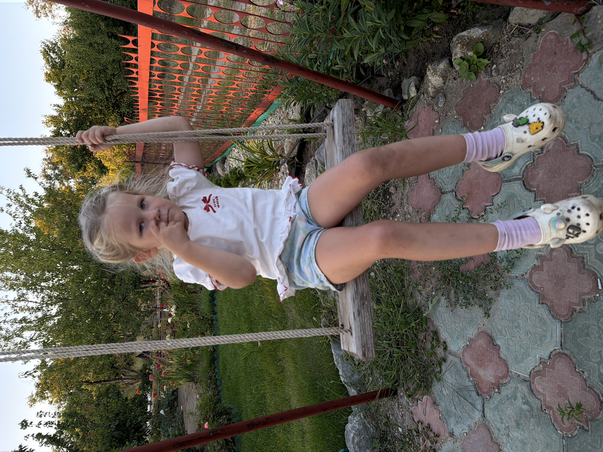 A young girl with blonde hair is sitting on a wooden swing in a garden. She is wearing a white t-shirt with a red bow graphic and light blue shorts. She has on white Crocs with charms and light purple socks. The girl is looking towards the camera with her fingers in her mouth, appearing thoughtful or a bit shy. She is holding onto the swing's rope.
The setting is a garden with green grass, trees, and some flowering plants. A red metal fence is visible in the background. The ground is paved with decorative stones. The lighting suggests it is daytime, possibly late afternoon given the warm tones.
There is text visible on the girl's t-shirt: "true match". The overall mood is peaceful and captures a moment of childhood in a natural setting. The location is Kamyshin, Russia.