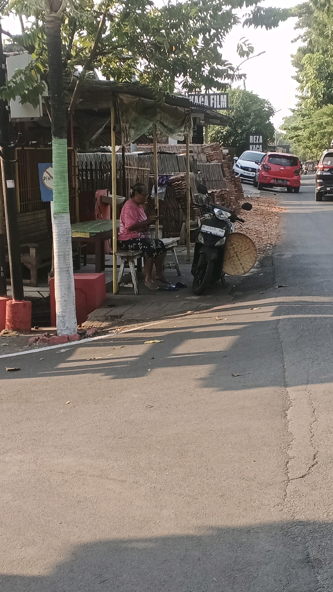 The image captures a scene on a street in Semarang, Indonesia, with a woman sitting on a low stool under a makeshift shelter. She is wearing a pink t-shirt and dark patterned pants and appears to be looking at her phone. A motorcycle is parked next to her, with a large, round woven mat leaning against it. Behind the woman and the motorcycle, stacks of what appear to be roofing tiles are visible, along with a sign that reads "KACA FILM" (meaning "Window Film" in Indonesian) and another sign with the name "REZA KACA." The shelter itself is constructed with a corrugated metal roof and tarp material. The street is paved, and trees line the right side of the road. The lighting suggests it is late afternoon, with long shadows cast on the ground. The overall impression is of a small roadside business or stall.