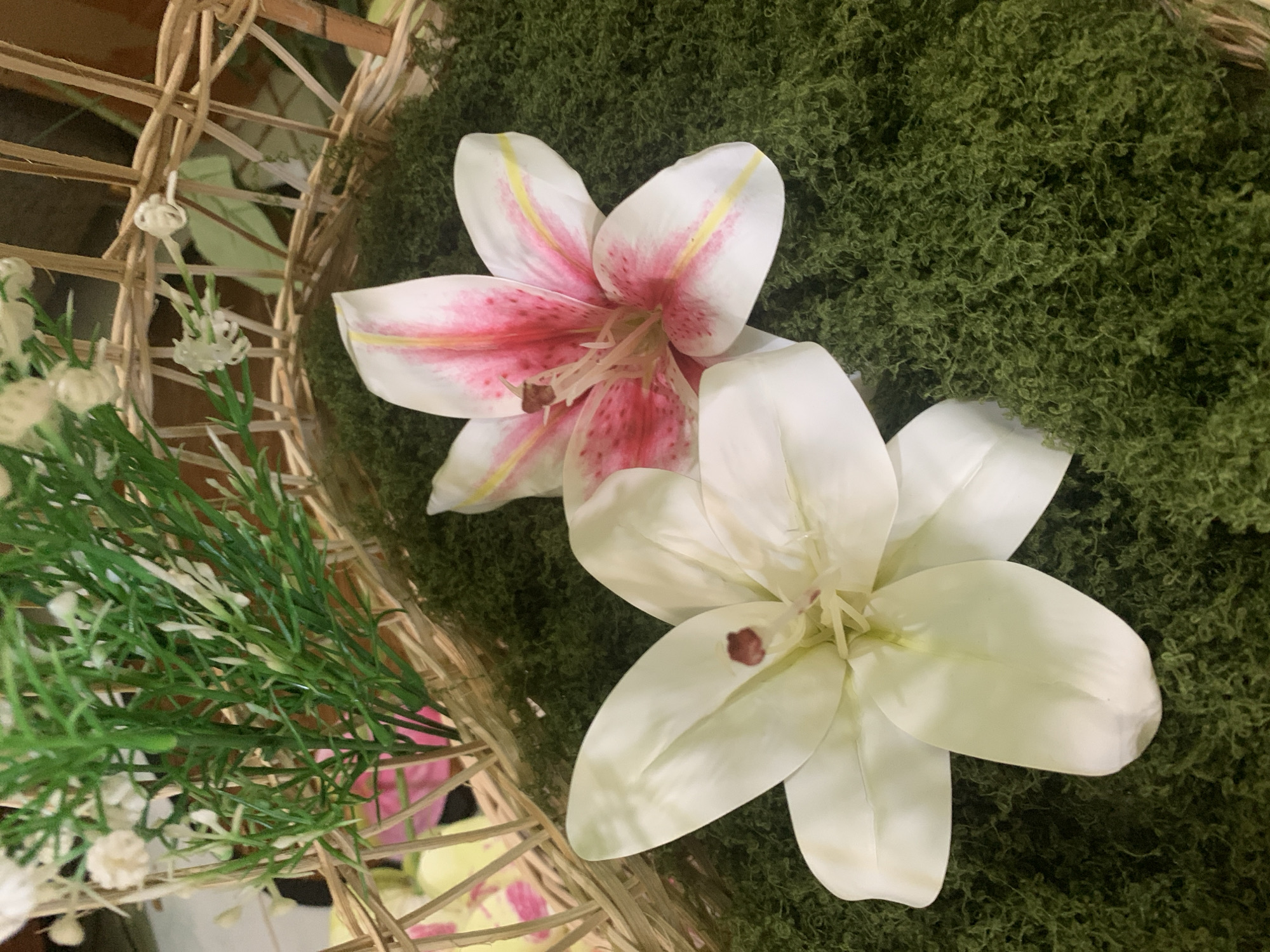 A close-up, overhead shot shows two artificial lilies, one white and one pink and white, resting on a bed of green moss within a woven basket. To the left, artificial greenery with tiny white flower buds extends upwards. The background consists of a blurred brown surface and the woven texture of the basket. The image is static and shows no people or activity. The location is unspecified in the image itself.