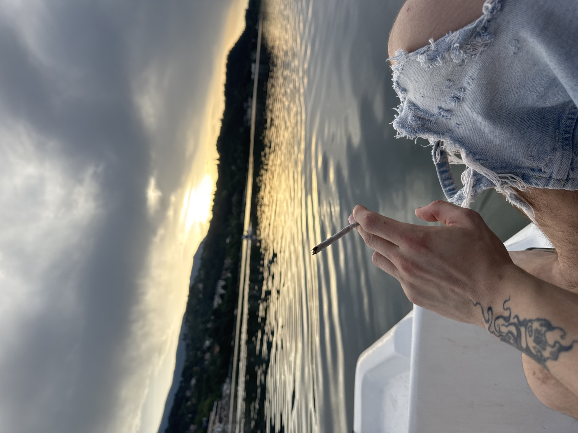 A person, partially visible from the chest down, sits in a white plastic boat on a body of water. The person is holding a lit cigarette in their right hand, which is positioned in the foreground. They are wearing distressed denim shorts. The background shows a landscape of hills covered in trees and buildings, reflecting on the rippling water. The sky is overcast with dramatic clouds, and a bright spot of sunlight is visible near the horizon, suggesting either sunrise or sunset. The location is identified as Pancharevo, Bulgaria.