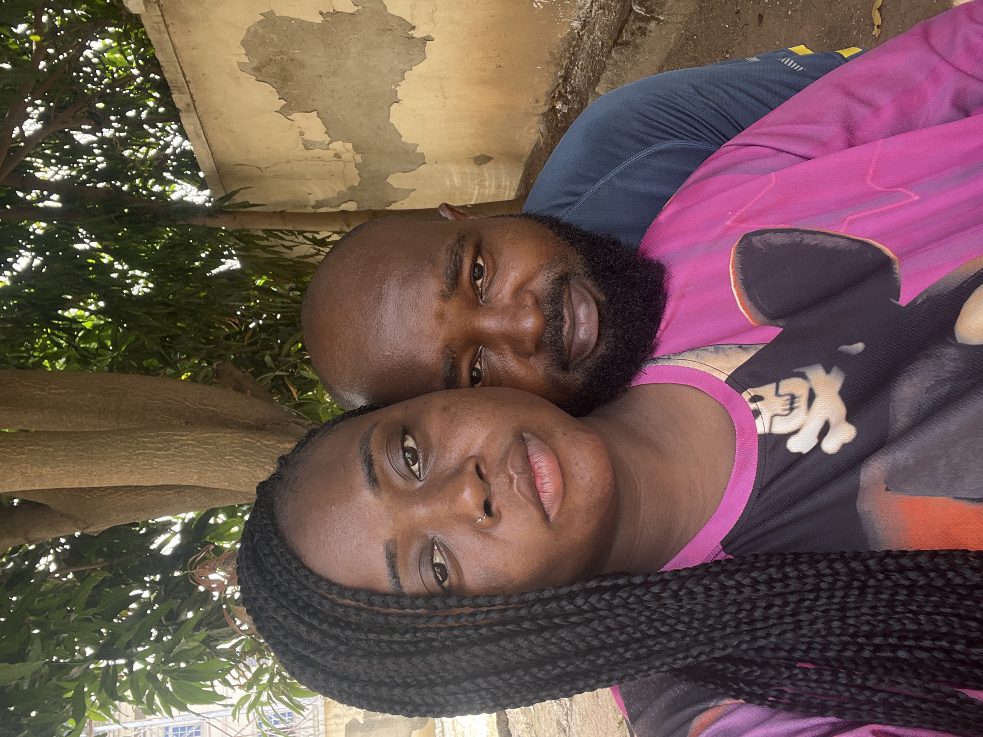 A close-up, portrait-oriented photo, taken outdoors in Kubwa, Nigeria, features a man and a woman, likely a couple, looking directly at the camera. The image is rotated counter-clockwise.
The woman is in the foreground, positioned to the lower right of the frame (when upright). She has dark skin, with long black braided hair falling over her shoulders. She wears a silver nose piercing and a multi-colored top, primarily pink and black, featuring what appears to be a stylized cartoon ear (possibly Mickey Mouse) and some white graphic elements resembling a skull and crossbones or a similar motif. Her expression is calm, with a slight, subtle smile.
Behind and slightly above her, the man is visible. He also has dark skin, a bald head, and a well-groomed full black beard. He wears a dark blue long-sleeved top with a yellow detail on the shoulder that is not clearly legible. He looks towards the camera with a relaxed expression.
The background behind them shows a mix of elements. To the upper right (when upright), there's a textured, light-colored wall or concrete structure with peeling paint and visible signs of wear. To the upper left and extending downwards on the left side, lush green tree foliage is visible, with bright sunlight dappling through the leaves, indicating a sunny daytime setting. A thick tree trunk is also visible behind the woman's head. The overall mood is relaxed and intimate. There is no discernible text on either person's clothing or in the background.
