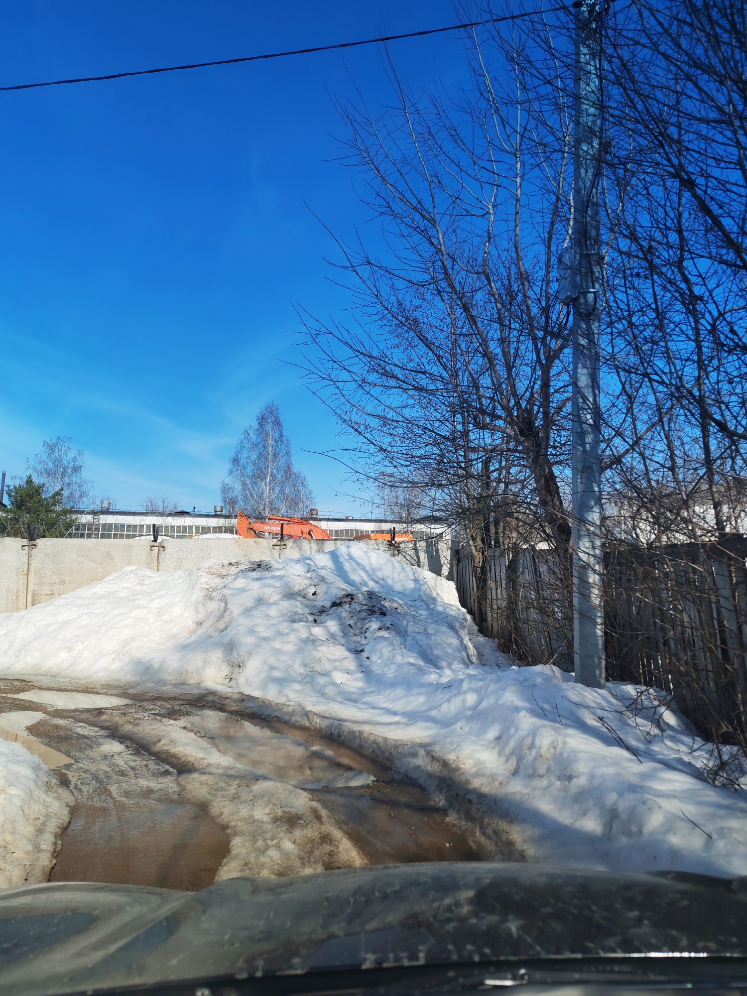 The image shows a winter scene, likely in a rural area, with a large pile of snow on the side of a partially snow-covered, muddy road. Tire tracks are visible in the mud. In the background is a grey concrete wall topped with barbed wire, behind which a long, low building with multiple windows is seen. An orange excavator is partially visible behind the snow pile. A telephone pole stands amidst bare trees to the right. The sky is clear and blue. No people are visible in the image. Without additional context, the specific location within Chaikovsky, Russia, cannot be verified.