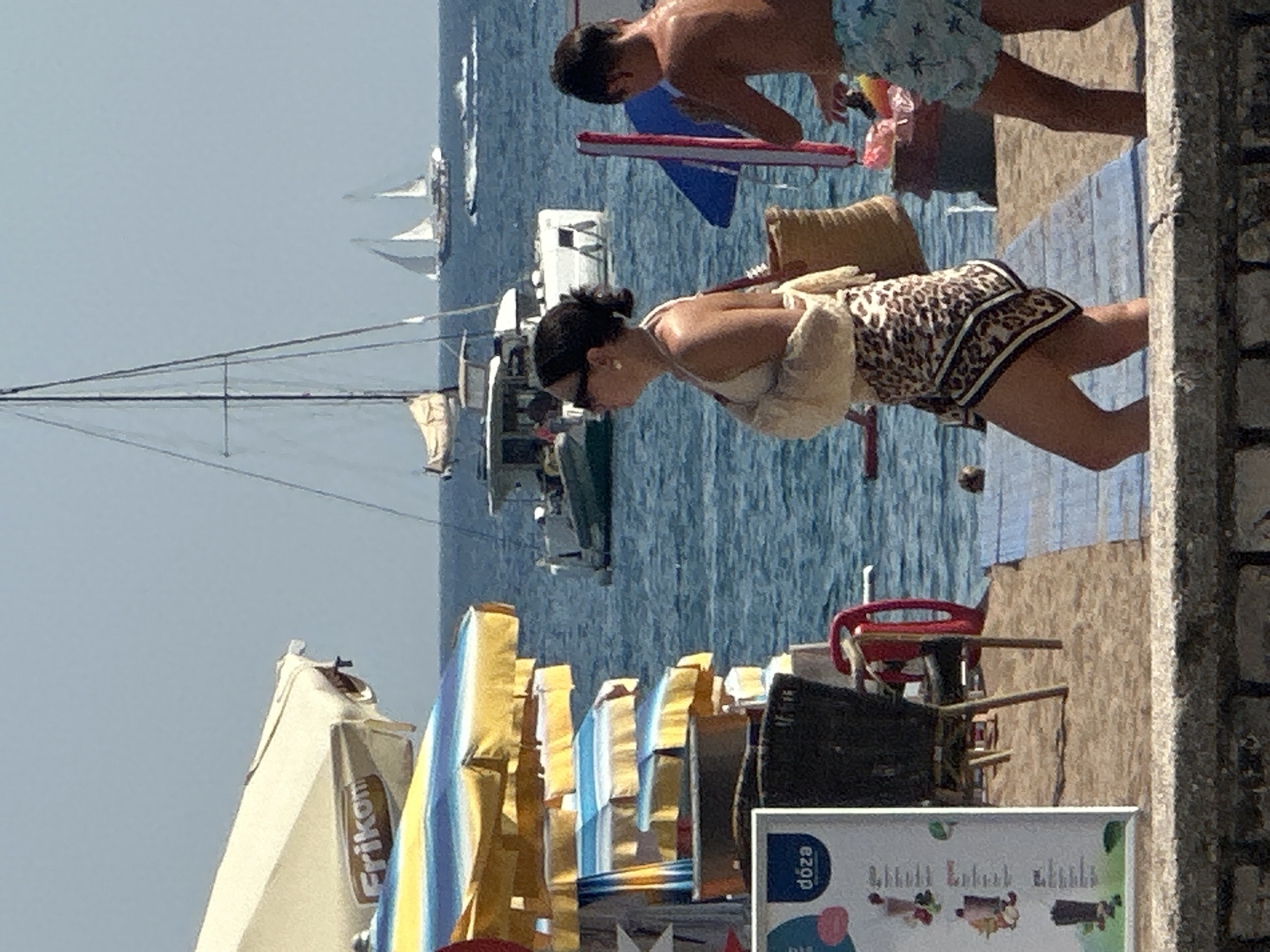 This image captures a beach scene in Boreti, Montenegro, on a sunny day. In the foreground, two people are visible from the waist down, facing away from the camera. One individual, wearing a leopard-print skirt and a light-colored top with a sling on their left arm, stands on a blue and white striped mat. To their right, another person, wearing blue patterned swim trunks, is also on the mat, appearing to reach for something. Behind them, several beach umbrellas, some stacked and others open, are set up on the sand. A menu board with "dóza" written on it is positioned to the left. The background features the blue sea with several sailboats. One large sailboat with white sails is prominent, with two smaller sailboats further in the distance. Two white motorboats are also visible on the water, one closer to the foreground. The sky is clear and blue.
