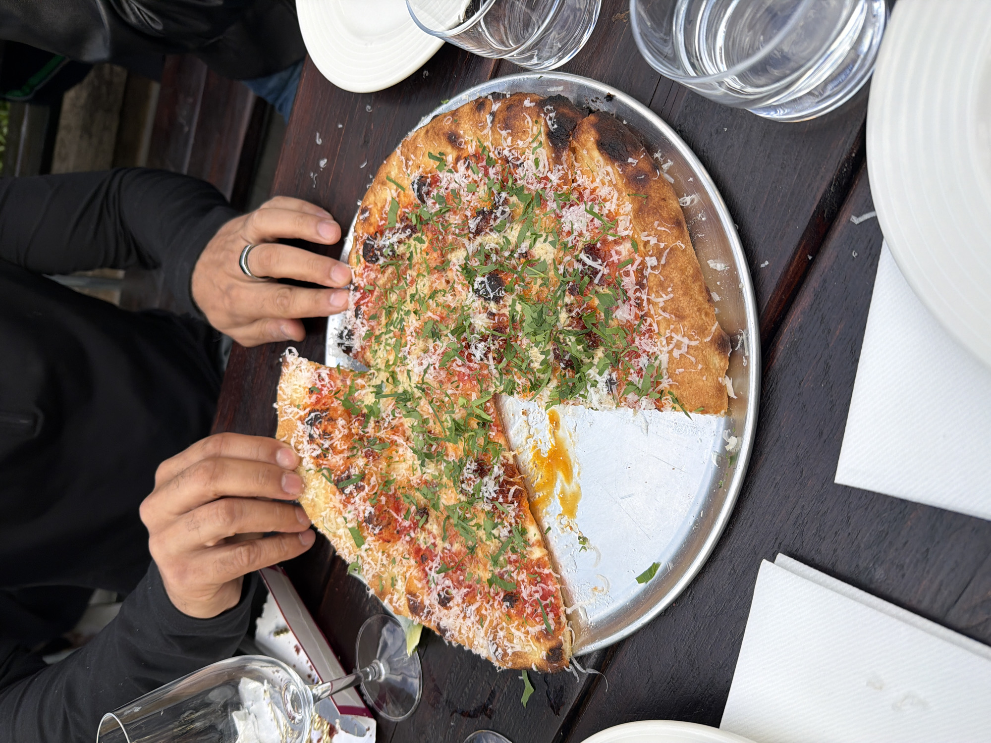 The image shows a person taking a slice of pizza from a metal tray on a dark wooden table. The pizza is round with a golden-brown crust, topped with red sauce, small dark-colored pieces, shredded white cheese, and green herbs. A portion of the pizza is missing from the tray. The person is wearing a long-sleeved black shirt and a silver ring is visible on their left hand. Beside the pizza, there are several clear glasses, white plates, and folded white napkins. The scene takes place outdoors, as suggested by the ambient lighting and the partial view of greenery in the background. There is no explicit evidence to confirm the location is in London, United Kingdom.