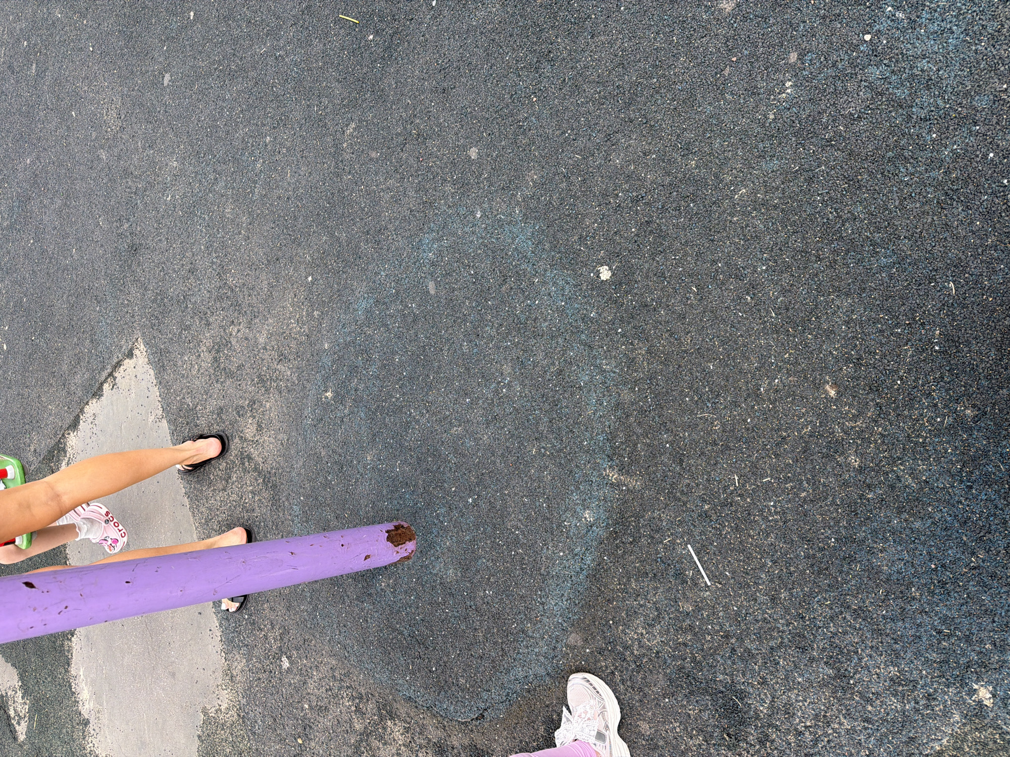 The image depicts a close-up, low-angle view of a purple pole standing vertically in the center of the frame, rooted in dark, textured ground cover. The ground appears to be a playground surface, possibly rubberized, with a circular pattern of lighter blue visible around the base of the pole. To the left, a person's lower legs are visible, wearing a white athletic shoe and pink leggings. To the right and slightly behind the pole, another person's legs are visible, wearing black flip-flops and light-colored shorts or a skirt. A hint of green object, possibly playground equipment, is visible in the background at the top edge of the frame. The scene suggests an outdoor recreational area, consistent with a playground in Lopatino, Russia, though no specific landmarks of Lopatino are identifiable within the frame. The lighting appears to be natural daylight, casting minimal shadows.