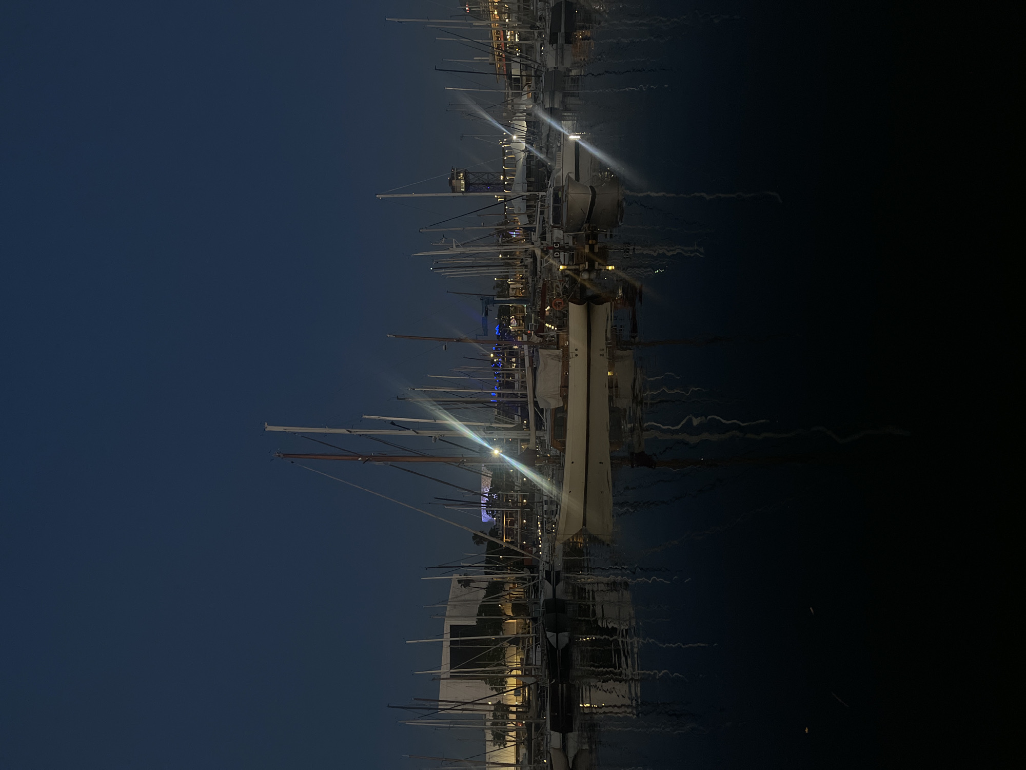 The image shows a nighttime scene in a marina with many sailboats. The sky is a dark blue, indicating it is dusk or night. The water is dark with reflections of the boats and lights. The scene seems to be in Barcelona, Spain. Several boats with masts and sails are visible, creating a dense network of lines against the sky. Light sources from the boats and possibly from structures on land reflect in the water. There are no visible people.