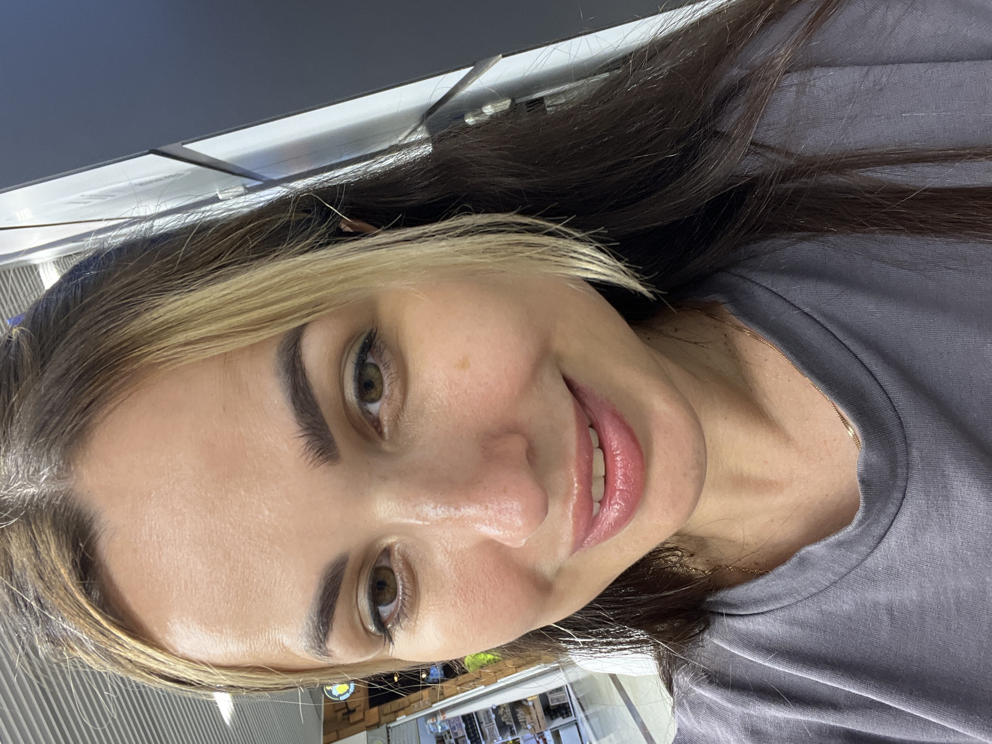 A woman is taking a selfie in a store. She has long dark hair with blonde highlights. She is wearing a gray t-shirt and a delicate gold necklace. Her skin appears dewy and she has on lip gloss. The background is out of focus, but it appears to be a well-lit indoor space with displays of merchandise.