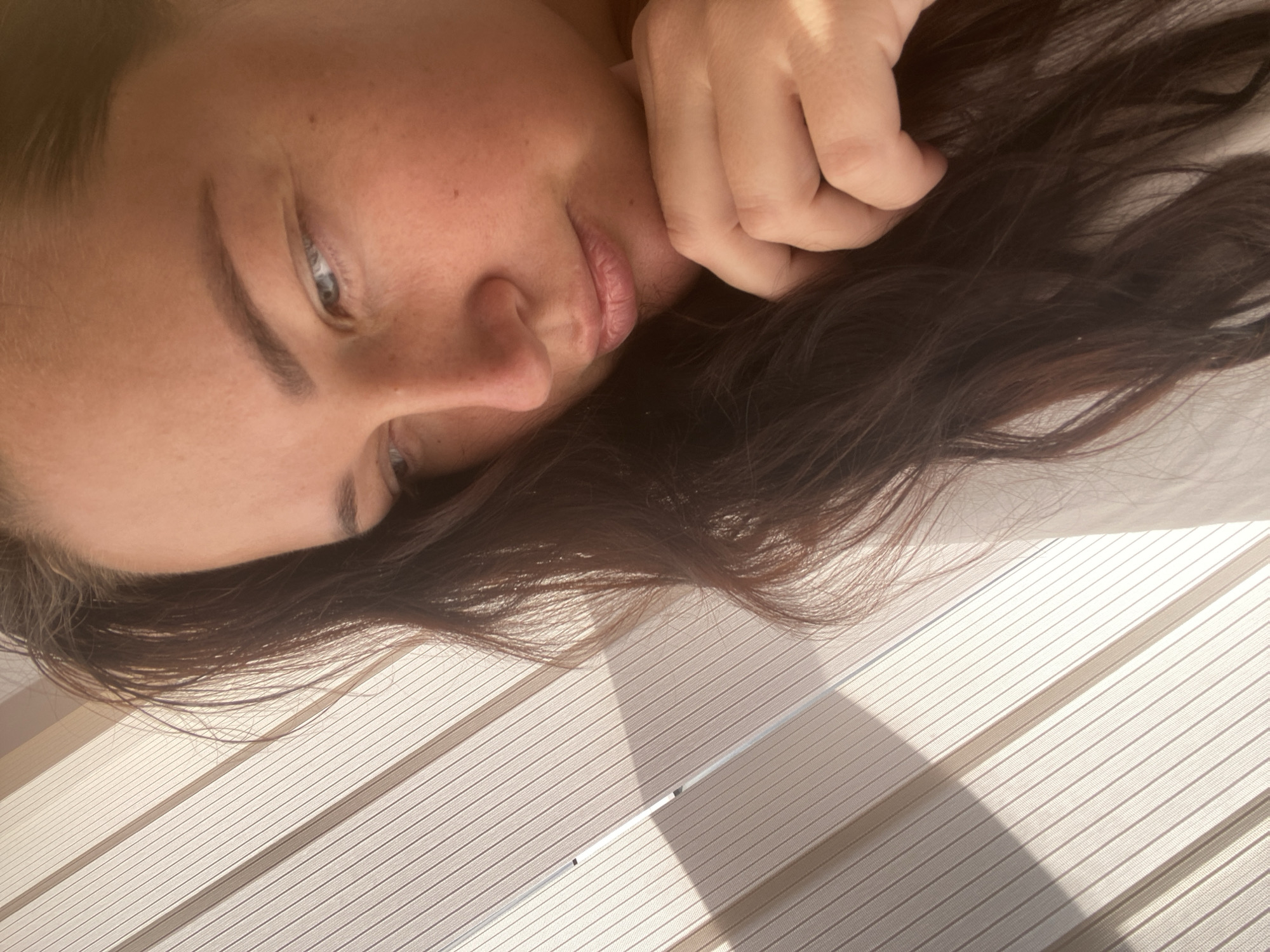 A close-up, shallow depth-of-field portrait shows a woman with long, dark brown hair resting her chin on her hand. She is looking downwards and to the left, with her eyes partially closed. The background is dominated by off-white blinds, with a shadow cast across them from an unseen light source. The lighting suggests it is daytime, likely late afternoon or early morning due to the warm tones. There are no discernible landmarks or features that place the image specifically in Sochi, Russia.