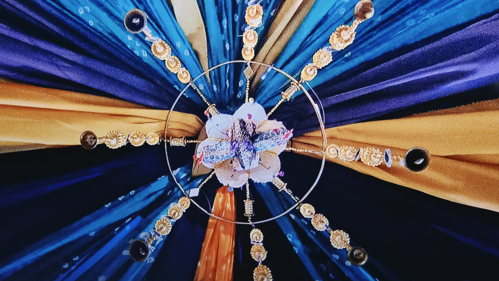 The image depicts a ceiling decoration, likely for an event or celebration. The decoration features alternating panels of golden yellow and teal/navy blue fabric emanating from a central point. A circular metal frame is suspended at the center, adorned with a floral arrangement and decorative chains with gold medallions and what appear to be dark-colored buttons or beads. The backdrop is a continuation of the radiating fabric panels. There are no people visible. The image provides no information that would locate this scene in the city of Kalipetrovo, Bulgaria.