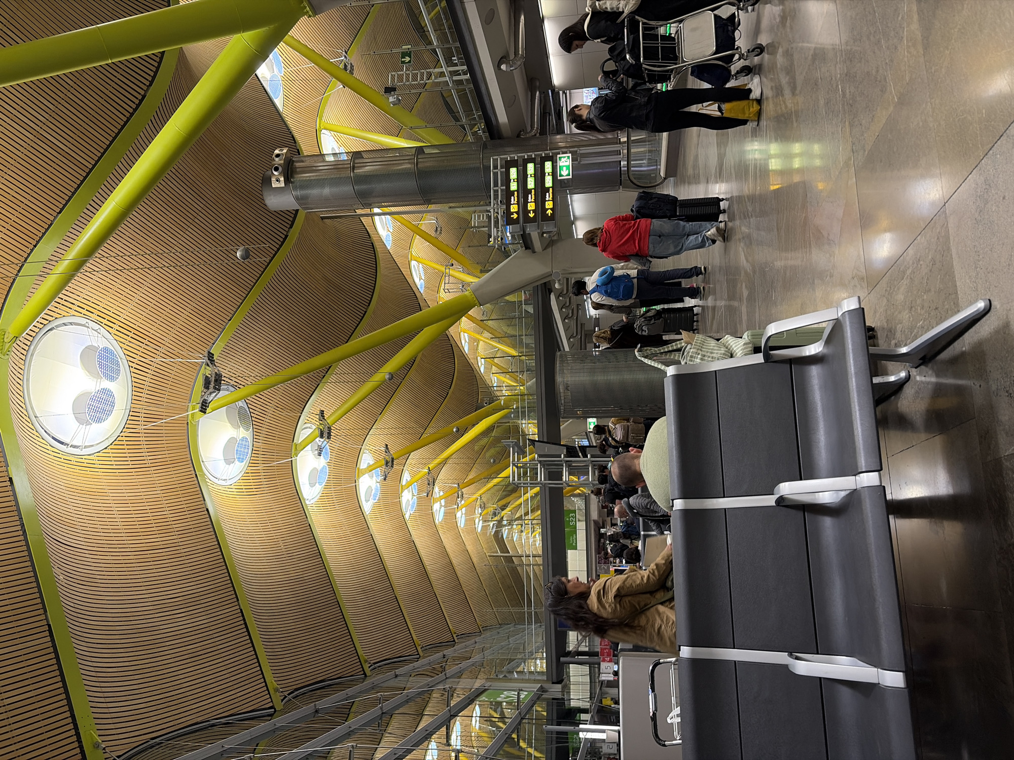 This image depicts the interior of an airport. The architecture features a high, curving ceiling made of horizontal wooden slats, supported by a network of bright yellow beams. Round skylights are embedded within the ceiling structure. The floor is a light grey, shiny material, likely stone or tile.
Several people are visible in the scene. Some are standing in line, and others are seated in rows of black plastic seating. Some people are pulling luggage.
Overhead, there's a large metallic sign with green text and symbols.