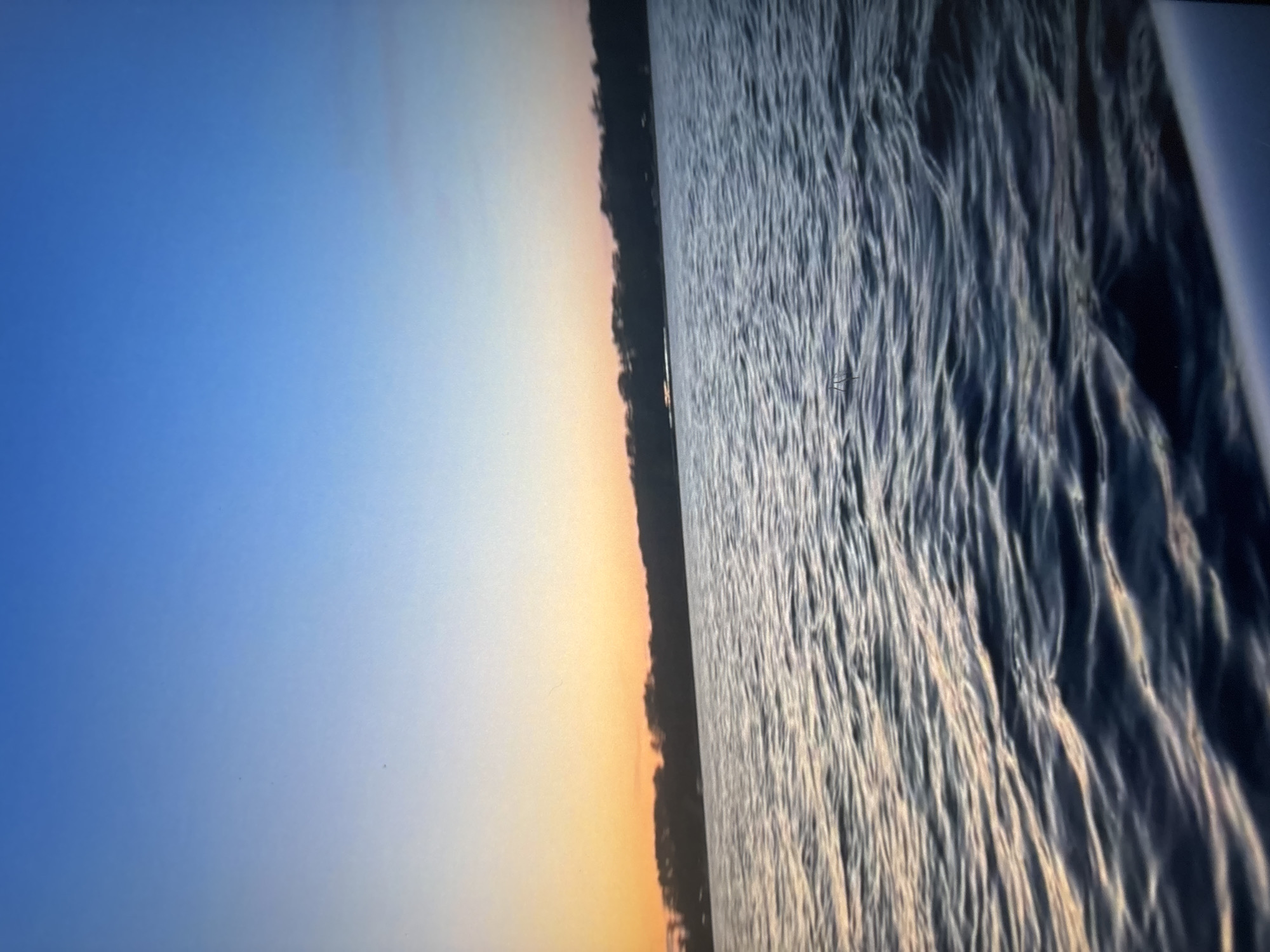 The image depicts a body of water with gentle ripples, reflecting the gradient of the sky. The sky transitions from a deep blue at the top to a soft orange and yellow hue near the horizon, suggesting either sunrise or sunset. A dark, tree-lined shore stretches across the middle ground of the image. The foreground shows a sliver of a white object, possibly the edge of a boat or a railing, hinting at the perspective of someone on the water. There are no people or animals visible in the scene. The scene is tranquil and natural, with no man-made structures clearly identifiable beyond the distant treeline. The location is not specified as Omsk, Russia, and there are no elements in the image that would definitively place it there.