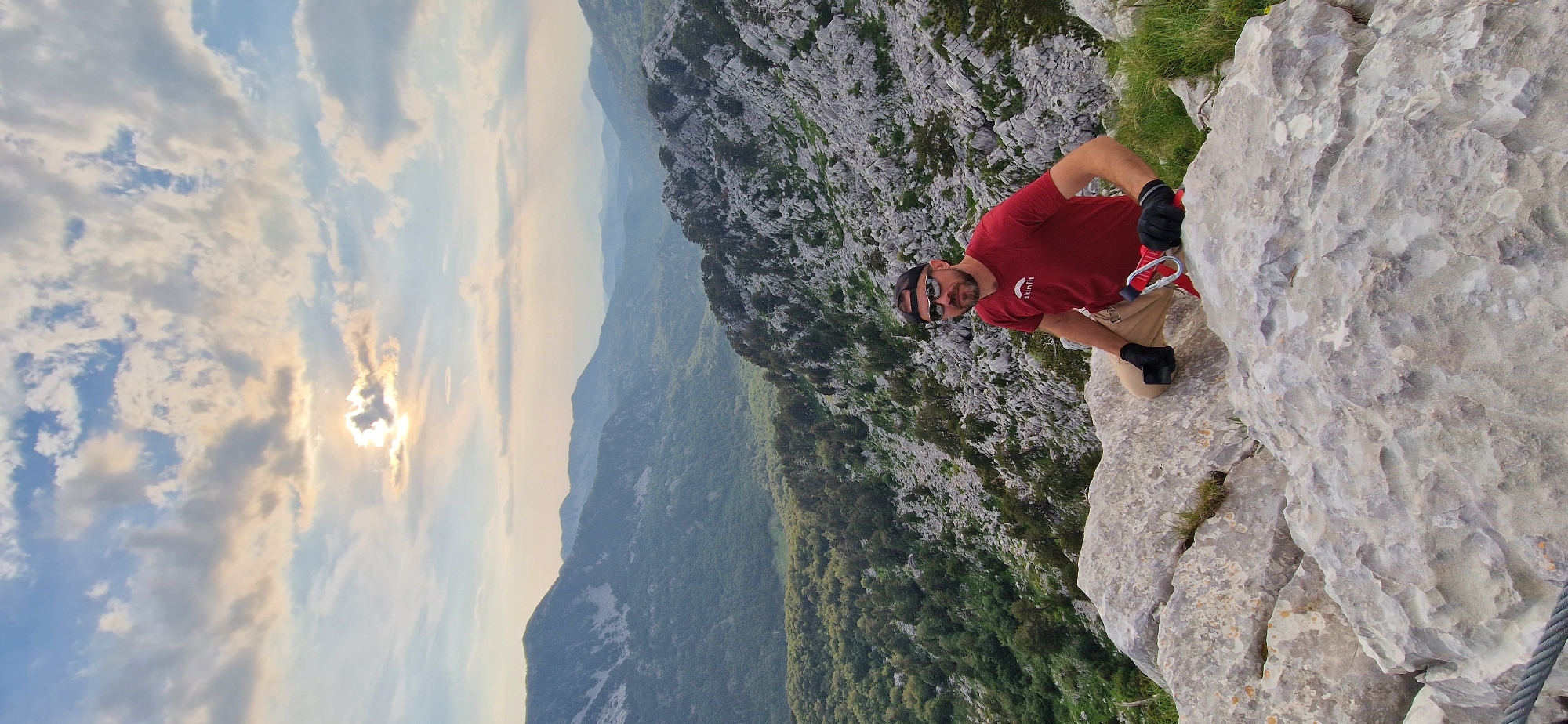 A man in a red t-shirt and beige pants is sitting on a large rock formation. He is wearing a black baseball cap and sunglasses. The man has a beard and is looking towards the camera. He is wearing black gloves, and a red climbing harness is attached to his pants. The man is positioned in the foreground, with a mountainous landscape in the background. The landscape consists of green forested valleys and rocky slopes under a cloudy sky. The location is identified as Kruševice, Montenegro. The scene appears to be a scenic overlook or a hiking spot in a mountainous region.