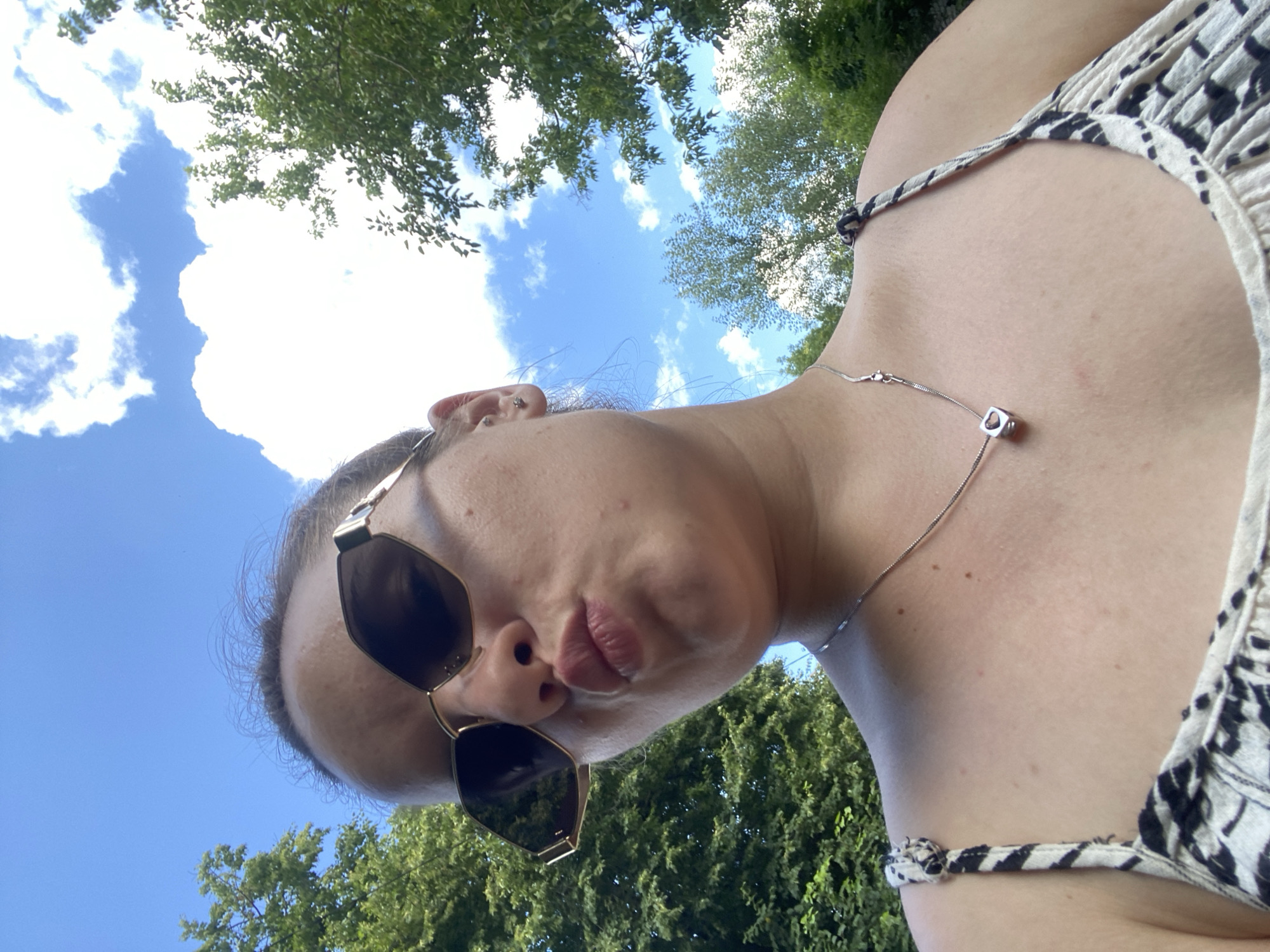 This is a selfie taken outdoors, with the camera angled upwards. The subject is a person wearing sunglasses and a patterned top. Their lips are pursed, suggesting a playful or attention-seeking pose. A thin necklace with a small, geometric pendant is visible around their neck.
The background consists of a bright blue sky with scattered white clouds and lush green trees. The foliage indicates a warm, sunny day, likely during the daytime. The camera angle and the framing of the shot suggest the person is looking up at the sky or a higher vantage point.
The image provides no visible text. The location context of Poltava, Ukraine, doesn't offer specific visual cues within the image itself, but places the scene geographically.