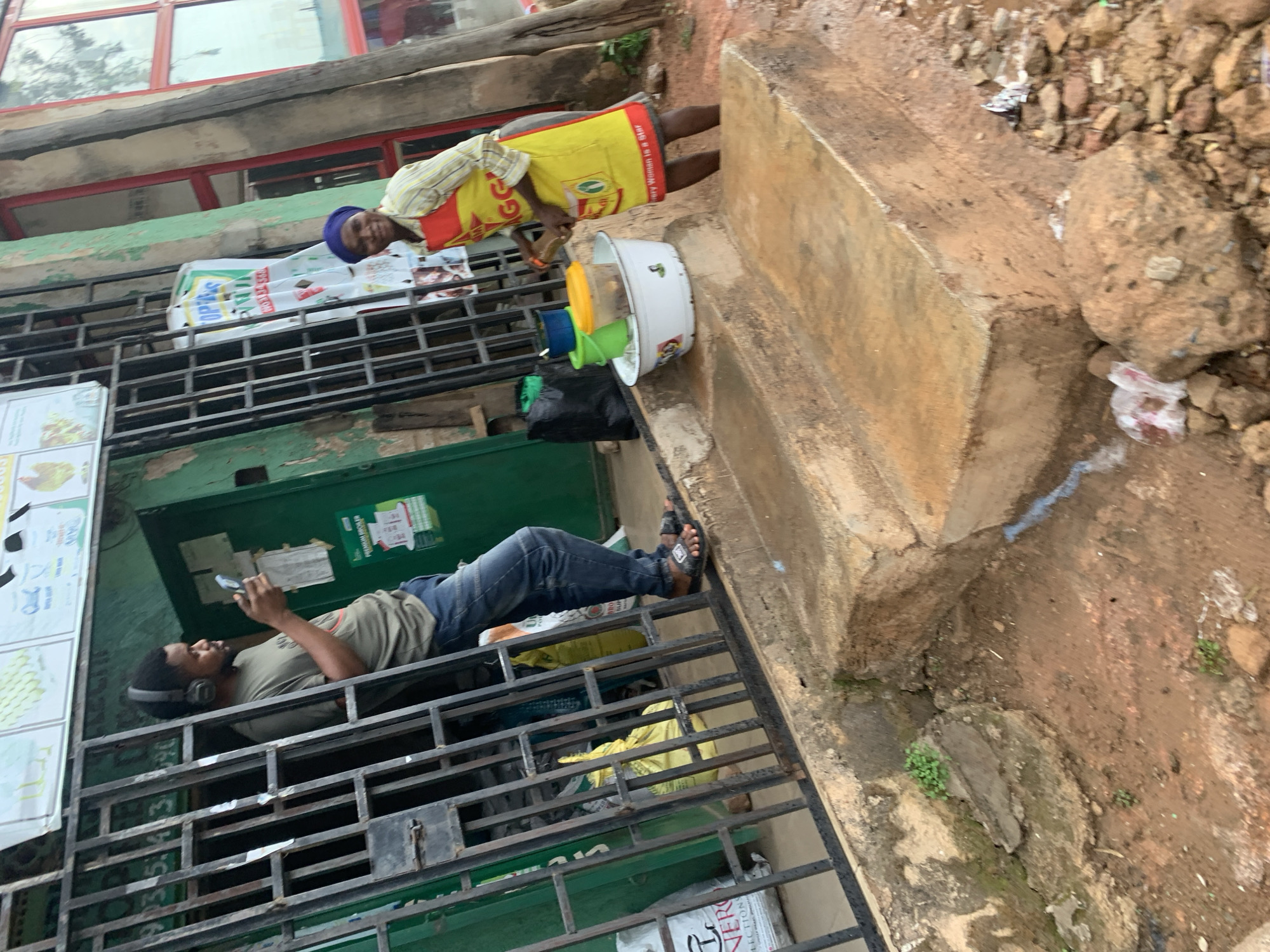 This image captures a street scene in Gura Top, Nigeria, featuring two individuals engaged in distinct activities.
On the right side of the frame, a person, possibly a woman, stands beside a concrete ledge. She is dressed in a yellow and red apron or vest over a striped shirt, with dark bottoms and a blue head covering. The apron bears a logo and faint text including "Every Woman is a Star." In front of her is a white bucket containing smaller, brightly colored containers (green, yellow, blue), suggesting a street vending setup. A black plastic bag rests on the ground nearby.
To the left, a man is lying on his back on a dark metal security grate or shutter, which covers the front of a storefront. He wears a grey t-shirt, blue jeans, and headphones. He is actively engaged with a smartphone in his hands. His feet, clad in black sandals, are propped up. Behind him is a building with a green-painted lower section and red-framed windows visible in the upper part of the background.
The ground around the concrete structure is unpaved, consisting of reddish-brown earth and loose rocks, with some small patches of green vegetation. Several posters and signs are affixed to the walls and the metal grate. One prominent sign behind the standing person displays "OPEX," "WHOLESALE," and "RICE." Another sign on the far left shows partial text, including "chicken."
The scene takes place during daylight hours, indicated by the natural illumination, and depicts a casual, everyday urban environment. The man appears relaxed, while the standing person seems to be attending to their wares.