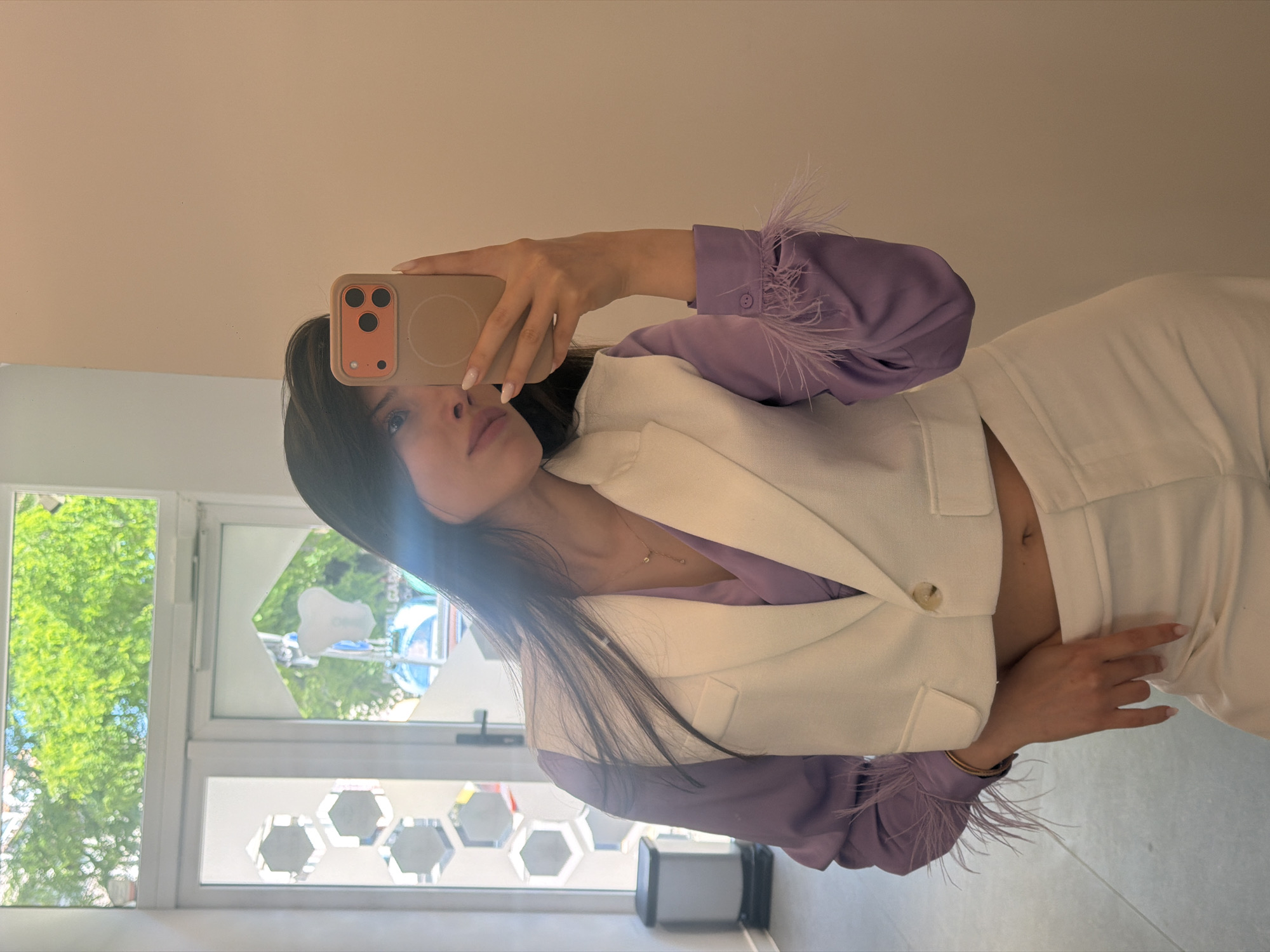 A young woman with dark hair takes a mirror selfie in an indoor location. She is wearing a lavender long-sleeved shirt with feather accents on the cuffs and a white cropped vest, paired with white pants. The background shows a doorway with a window, and through the window, green trees are visible. A trash can is to the left of the doorway. The wall behind her is a neutral beige color.