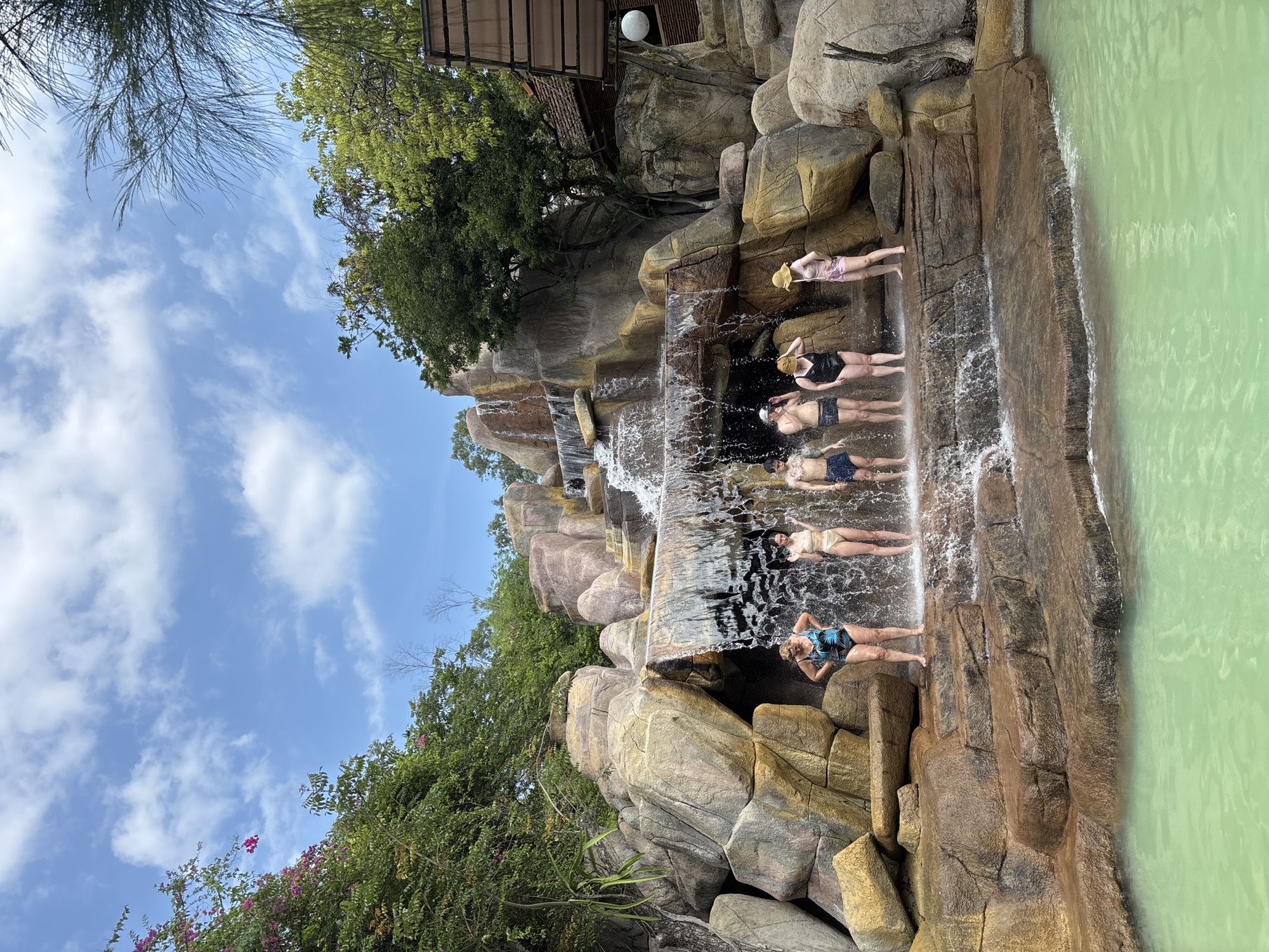 The image depicts a group of six individuals standing on a multi-tiered rock formation with a waterfall cascading down in front of them. The individuals, dressed in swimwear, are posed as if enjoying the water. The rock formation appears to be a man-made structure designed to resemble a natural waterfall. In the foreground, there is a pool of light green water. The background features lush green trees and a blue sky with scattered clouds, suggesting an outdoor setting. Part of a building with a thatched roof is visible on the right side of the image. The scene is likely located in an amusement park or resort in Tay Nha Trang Ward, Vietnam, given the presence of artificial rockwork and a pool.