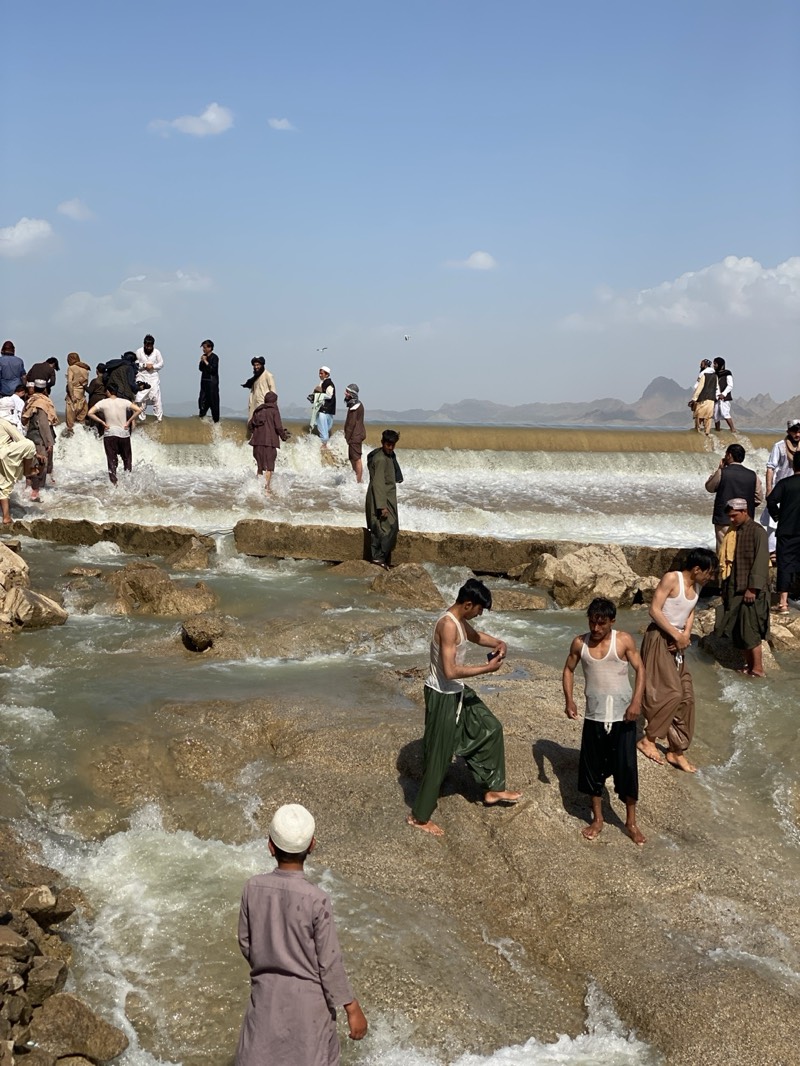 The image depicts a large group of Afghan men and boys, likely in the city of Kim, Afghanistan, gathered at a dam or weir. The men are of varying ages, dressed in traditional Afghan clothing, and some are barefoot or partially submerged in the flowing water. A few individuals are standing on the edge of the dam as the water cascades over, while others are wading in the stream below. One man is looking at his phone. The sky is light blue with a few sparse clouds. The landscape is rugged and hilly, with distant mountains visible. The overall scene suggests a recreational or social gathering in a natural setting.