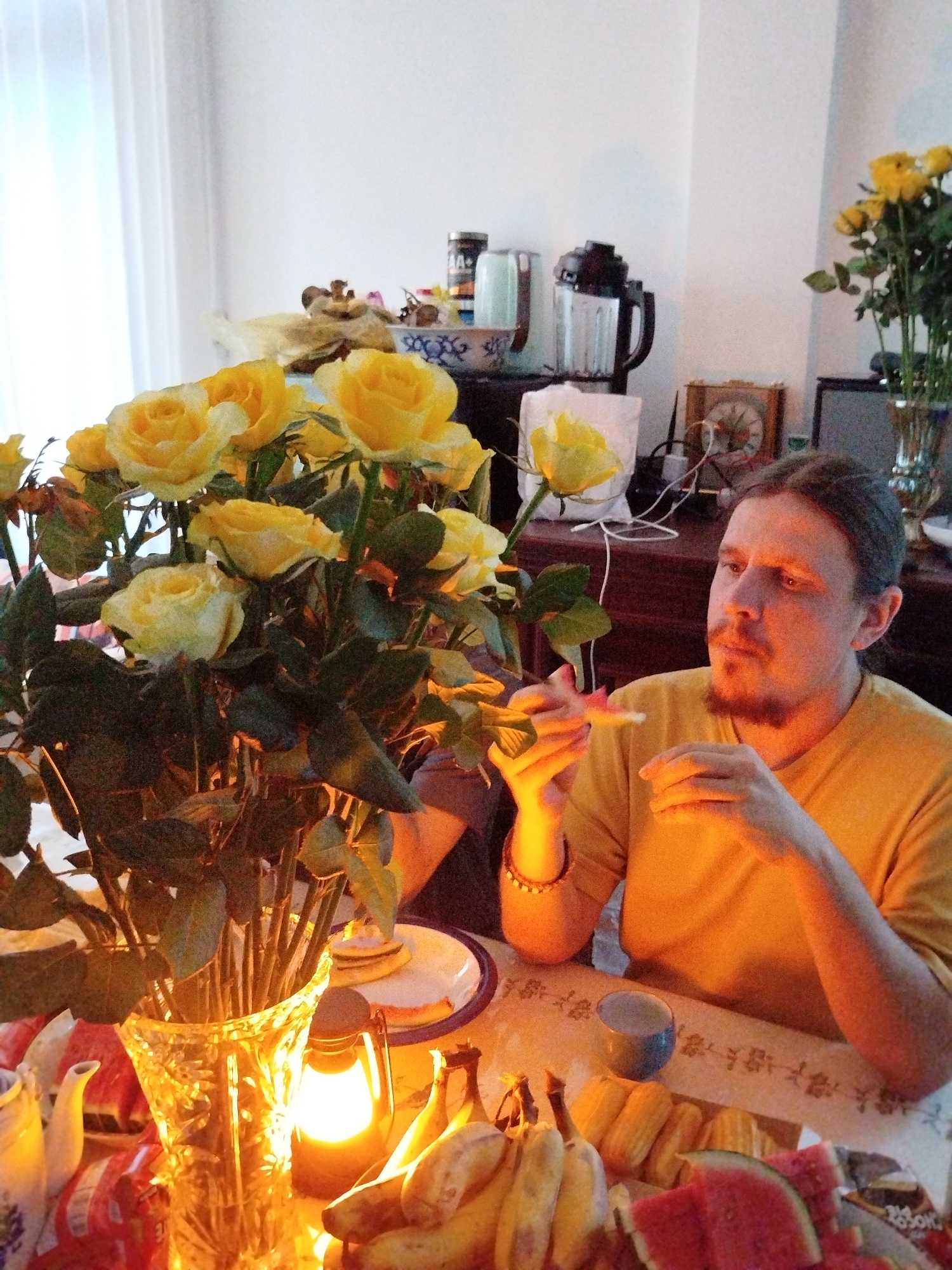 In an indoor setting, a man with long hair, a light complexion, and a reddish-brown beard is seated at a table. He wears a yellow short-sleeved shirt and a bracelet. The table is laden with a variety of items: a large bouquet of yellow roses in a crystal vase, bananas, slices of watermelon, and a small blue cup. A gas lantern provides illumination. A slice of watermelon is held in the man's right hand, close to his mouth. A cake sits on a plate behind the roses. Behind the man, a dark wooden dresser holds a blender, a clock, and another vase of yellow roses. A white curtain hangs next to the dresser. The scene is brightly lit, and the location is indeterminate. There is no identifiable evidence to suggest the setting is in Lạc Dương, Vietnam.