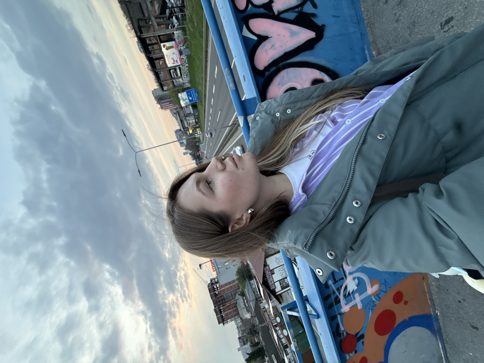 A young woman with light brown hair and fair skin is leaning on a blue metal bridge railing, wearing a grey-green jacket over a white and purple striped shirt. The railing is decorated with graffiti, including pink hearts and other colorful designs. Beyond the bridge, there is a view of Vozdovac Urban Municipality, Serbia, with buildings, roads, and a cloudy sky visible in the background.