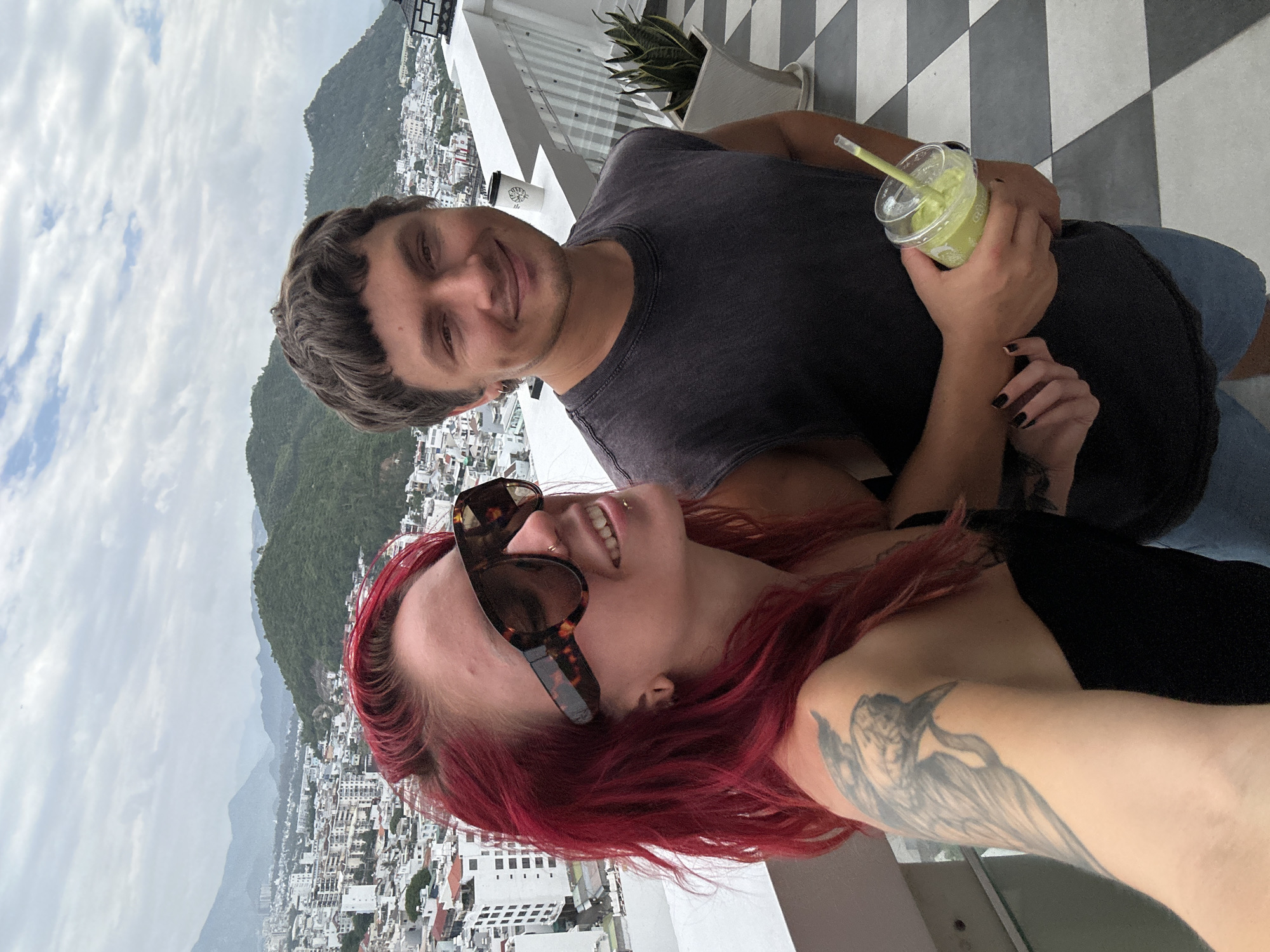A young couple poses for a selfie on a balcony overlooking the city of Nha Trang, Vietnam. The woman on the left has bright red hair and is wearing large tortoiseshell sunglasses. She has a tattoo on her left arm and shoulder. The man on the right has short brown hair and is wearing a dark grey sleeveless shirt. He is holding a green smoothie in a clear plastic cup with a straw. In the background, the city of Nha Trang stretches out to the sea, with buildings climbing up the surrounding hills. The sky is cloudy. The balcony floor has a black and white checkered pattern. A white paper cup is visible on the balcony railing.