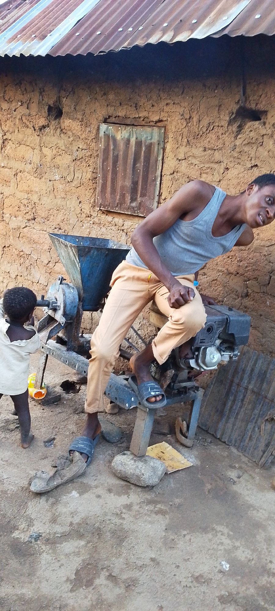 A young Black man in a gray tank top and tan pants is seated on a machine with a blue hopper and a metal engine, with his left foot resting on a support. A small Black child in a white shirt is standing to the left of the machine, facing it. The scene takes place outdoors in front of a mud brick building with a corrugated metal roof. The ground is dirt and gravel. The image appears to have been taken in Kamantan, Nigeria.