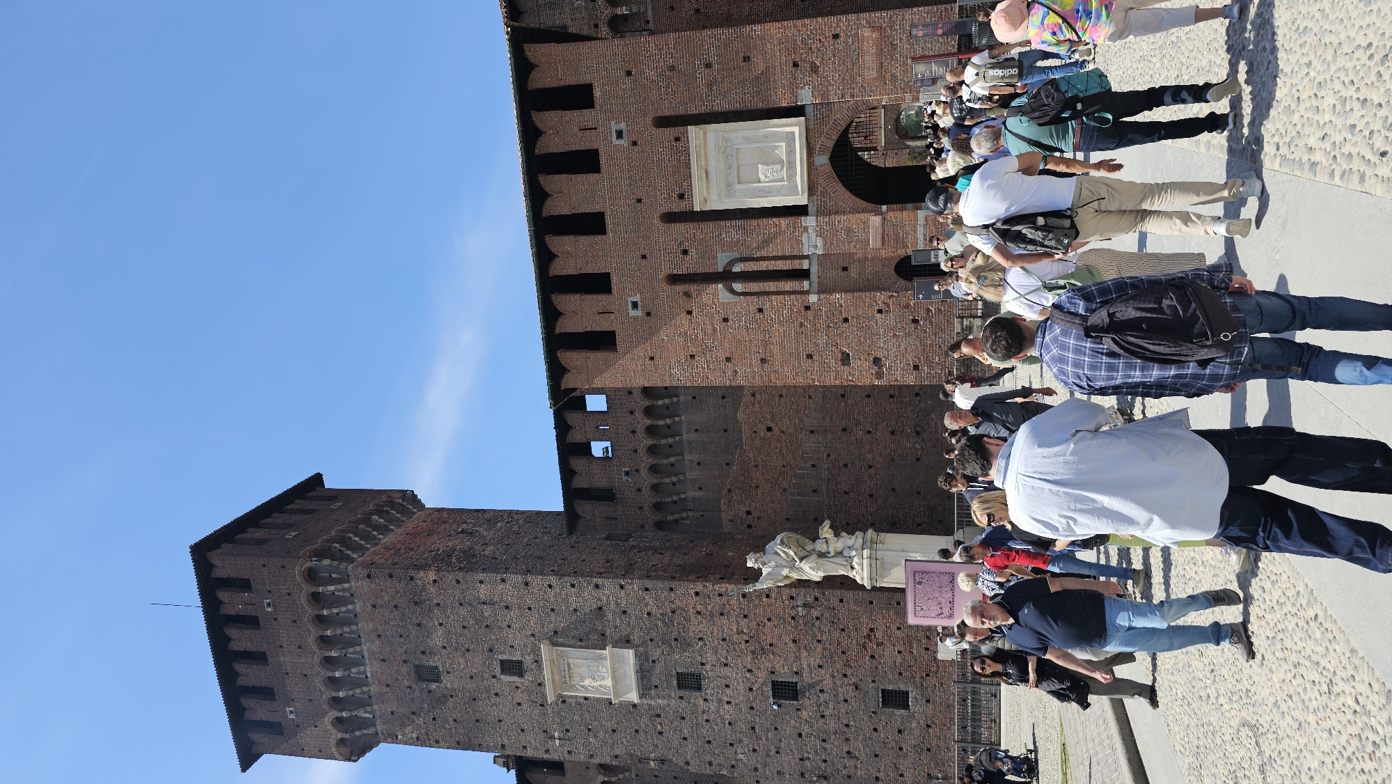 The image shows a low-angle view of the Sforza Castle (Castello Sforzesco) in Milan, Italy. The brick facade of the castle is visible, with towers extending upwards. People are milling around the base of the castle, some standing and others walking. A few are dressed in casual clothing. The sky above is clear and blue. A stone statue and a sign are also located near the castle entrance. The ground is paved with cobblestones.