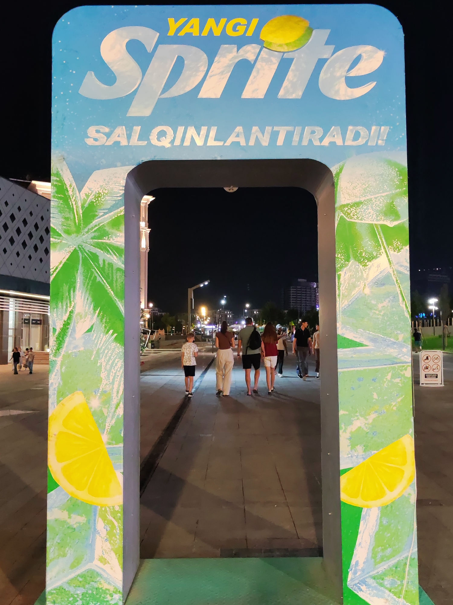 This is a night-time, eye-level, medium shot of an outdoor Sprite branded archway in Tashkent, Uzbekistan. The archway is decorated with large lemons and palm leaf graphics against a sky-blue background. The words "YANGI Sprite" and "SALQINLANTRADI!!" are prominently displayed in large white letters. Through the archway, a group of people are walking away from the camera on a paved walkway. The walkway is illuminated by streetlights and the lights from nearby buildings. The environment appears to be a public space, possibly a park or a pedestrian zone. The overall atmosphere is festive and inviting.