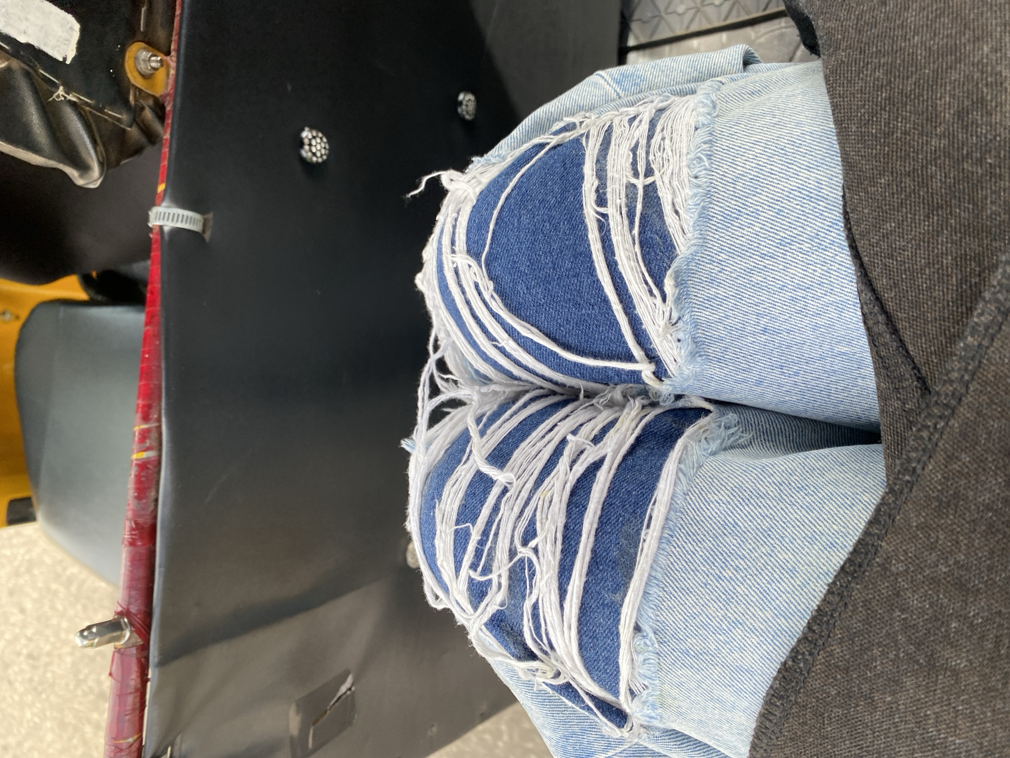 The image shows a close-up view of a person's legs wearing distressed denim jeans with rips and frayed white threads. The person is seated on what appears to be the passenger seat of a motor vehicle, likely an auto-rickshaw or Keke Napep, indicated by the black plastic body and a red-painted bar with a metal protrusion visible at the top. A yellow seat is partially visible behind the person. The background is out of focus, showing a greyish surface that could be a road or pavement. The scene is captured from a top-down perspective. The location is specified as Barakin Lerawi, Nigeria.