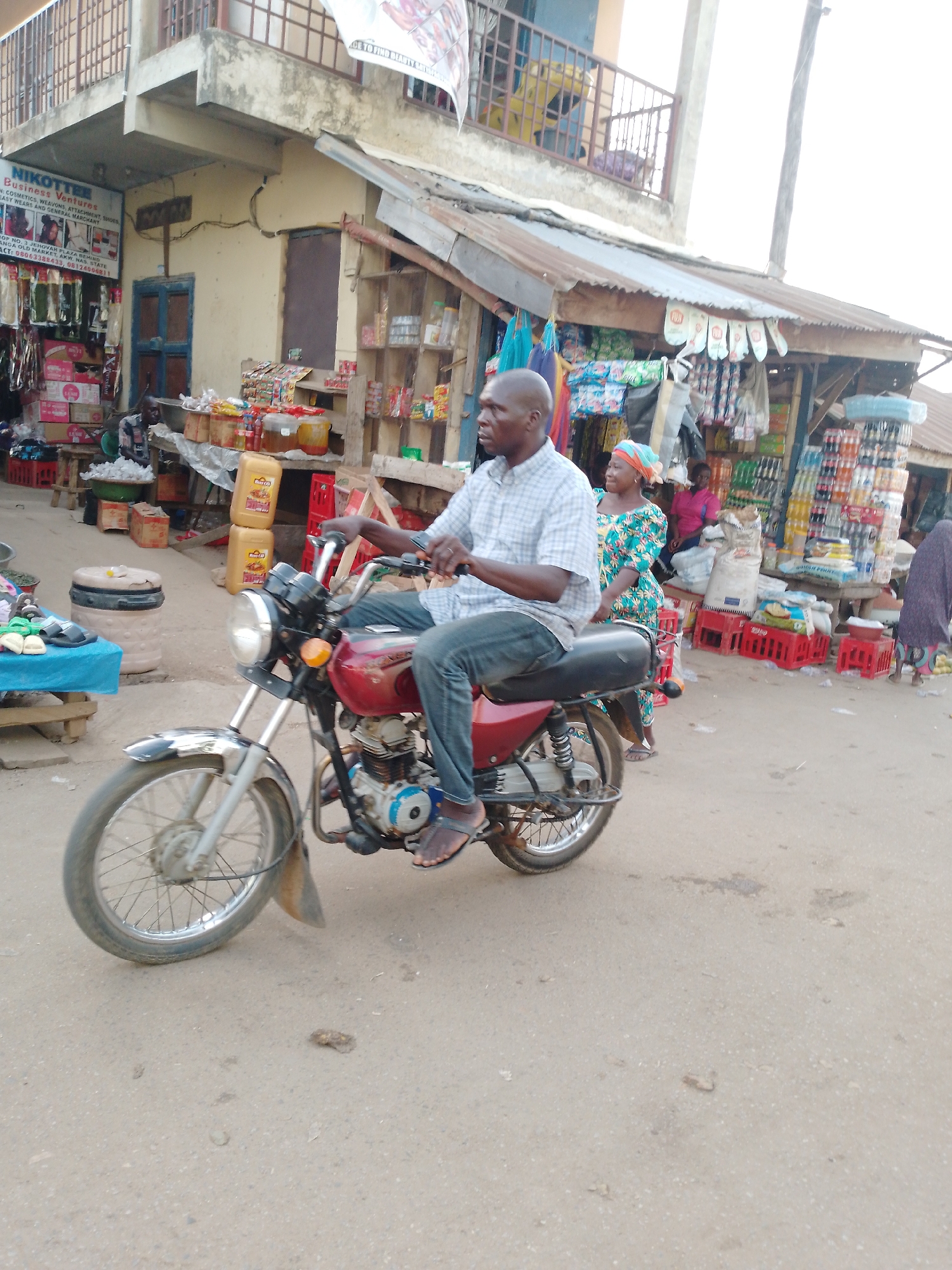 This daytime photograph captures a vibrant street market scene in Akwanga, Nasarawa State, Nigeria. In the foreground, a bald man in a plaid shirt and jeans sits on a red motorcycle, his feet in sandals touching the dusty ground, looking towards the left.
The background reveals a bustling commercial area with multi-story buildings and makeshift stalls. On the left, a building with a balcony houses "NIKOTTEE Business Ventures," identified by a prominent sign. The sign details the business offerings: "COSMETICS, WEAVONS, ATTACHMENT, SHOES, EASY WEARS AND GENERAL MARCHANT." It also provides location cues: "SHOP NO. 3 JEHOVAK PLAZA BEHIND ANGA OLD MARKET, AKW. NAS. STATE," and contact numbers: "08063388433, 08124006811." Below the sign, various packaged goods and slippers are displayed.
To the right, market stalls with corrugated metal roofs are packed with merchandise. Yellow jerry cans of oil are prominently stacked alongside various packaged foods, drinks in red crates, and other general goods. A woman wearing a patterned blue and green dress and an orange headscarf stands near the motorcycle, looking to the right. Further back, other individuals are visible amidst the market activity. A banner, partially visible in the upper right, includes the text "CE TO FIND BEAUTY STY." The scene suggests a busy local market on a clear day.