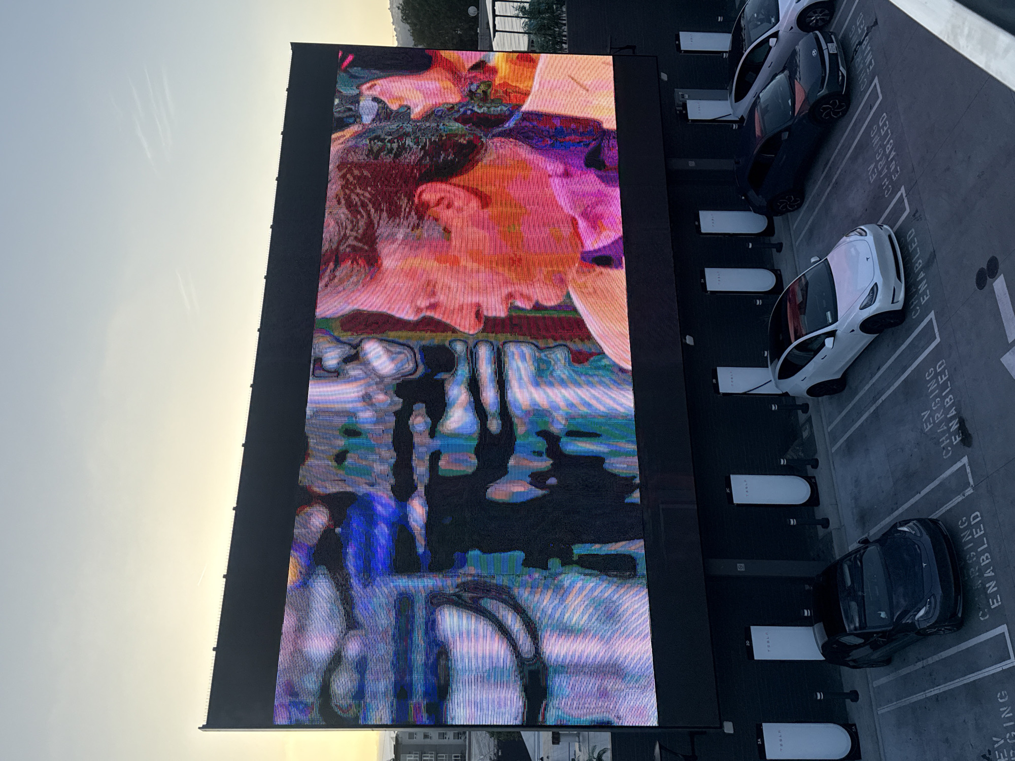 A large LED screen displays an image of two faces, heavily distorted with digital effects, and stands next to a Tesla charging station in West Hollywood, United States of America. Several Tesla vehicles are parked in charging bays, marked as "EV CHARGING ENABLED." The image is taken from an elevated angle, showing the screen, vehicles, charging stations, and a glimpse of the city skyline in the background. The sky is visible on the left side of the image.