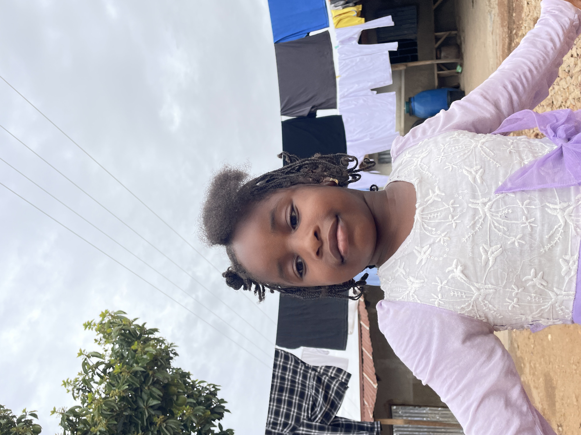 A young Black girl with braided hair stands outdoors in Jos, Nigeria. She wears a white dress with floral embroidery, long lilac sleeves, and a lilac bow. Laundry hangs on a clothesline behind her: blue, yellow, black, and white shirts and a black-and-white plaid garment. Power lines stretch across the overcast sky above. A green tree grows to the left, while a building with a brown roof and blue water container are in the background to the right.