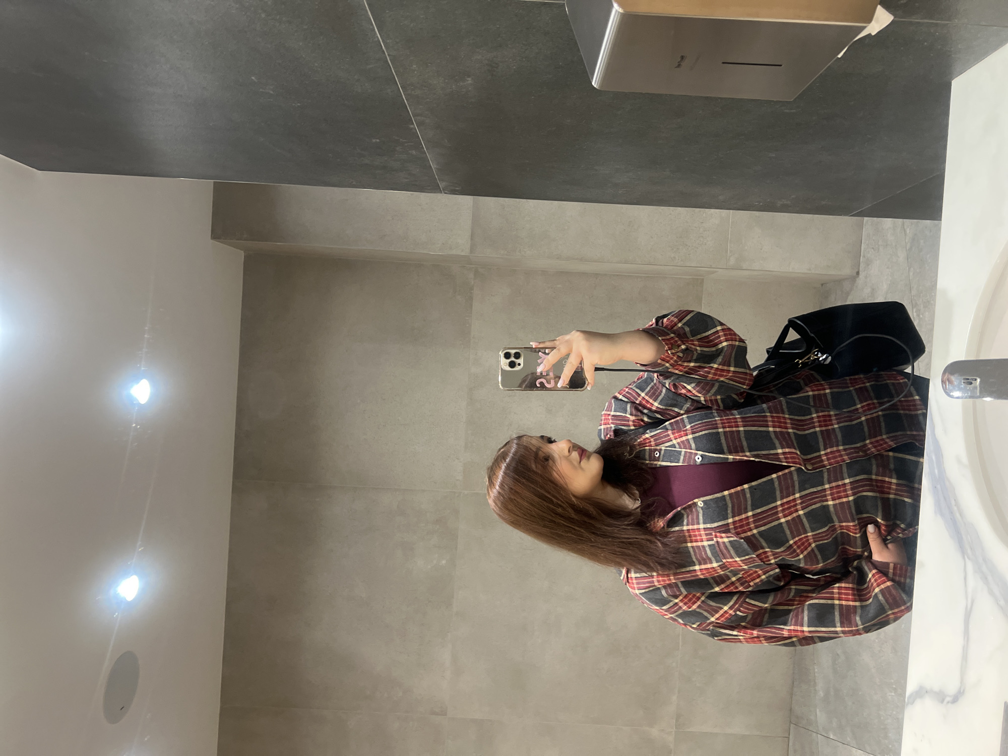 The image depicts a woman taking a selfie in what appears to be a modern restroom. The environment is characterized by a gray color scheme, featuring smooth, tiled walls and a darker, textured ceiling. To the right, a marble countertop is visible, alongside a sink and its faucet. There is also a black bag, likely a purse or tote, resting on the countertop. The woman is dressed in a plaid shirt and a dark top underneath. She is holding a cell phone, taking the photo of herself. Without further information, it is impossible to determine the exact location within Yerevan, Armenia.