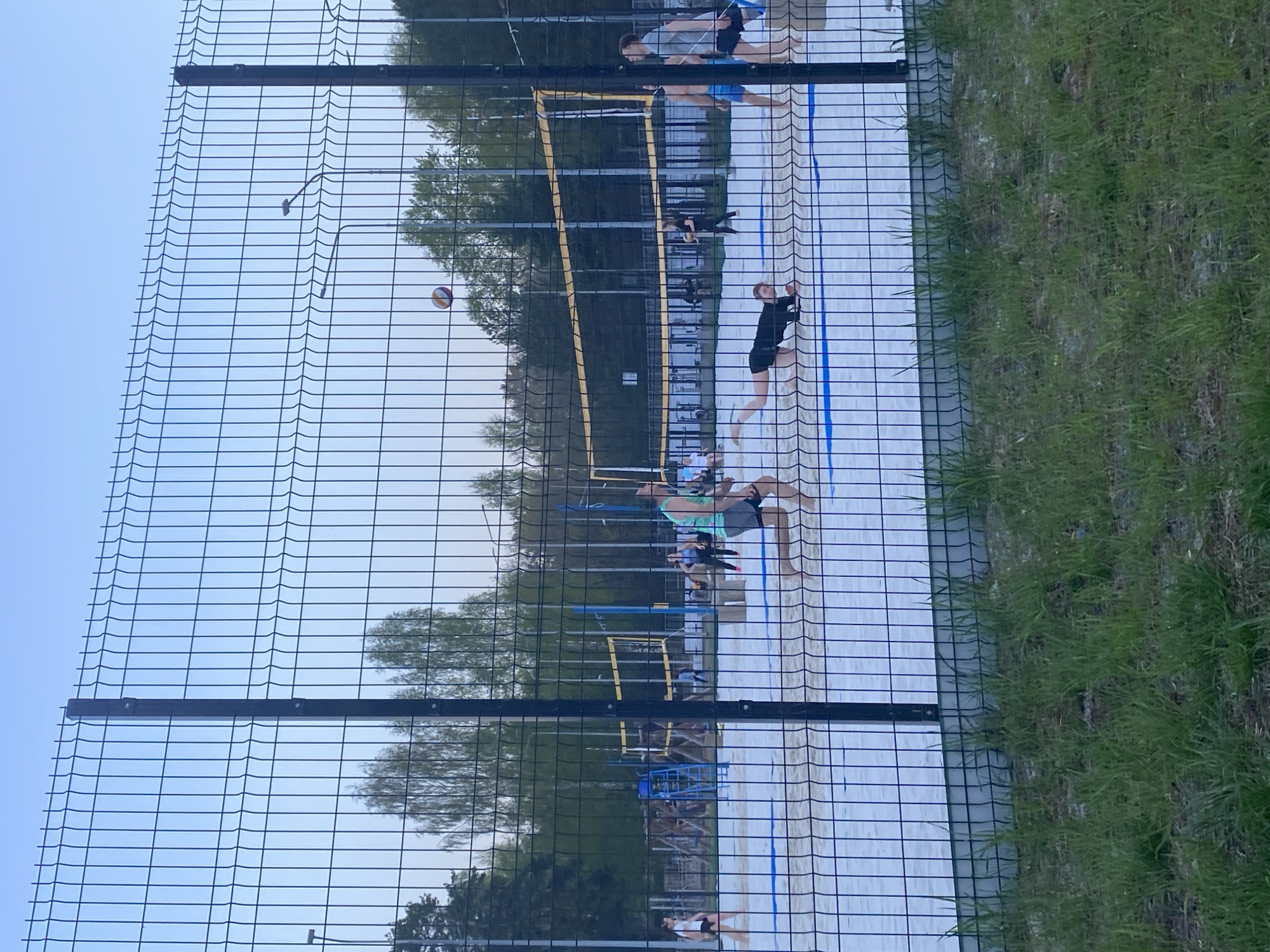 A photograph shows a beach volleyball game in Chaikovsky, Russia, as seen through a chain-link fence. The volleyball is in mid-air above the net. Several players are on the white sand court, some in action, others watching. Trees are visible behind the volleyball court. A grassy area borders the volleyball court.
