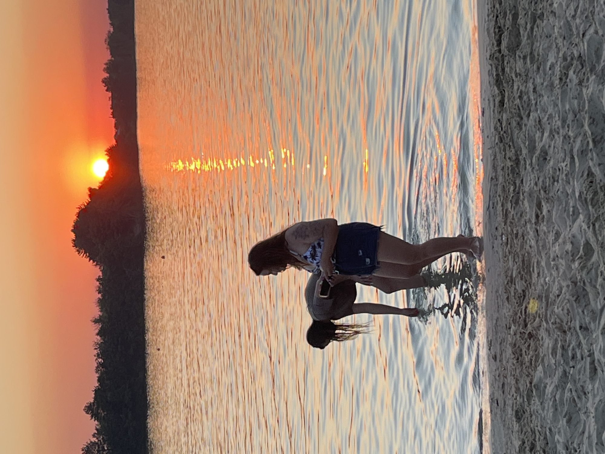 The image captures a serene sunset scene at a lake in Bezliudivka, Ukraine. The sky is ablaze with hues of orange and yellow, reflecting on the calm water. Two individuals are present near the shoreline. One person, a woman, stands facing right, holding a mobile phone in her hand. She is wearing a floral print top and denim shorts. Another person, with long dark hair, is bent over in the water, facing away from the camera. They appear to be looking for something in the shallows. The foreground shows a sandy beach, with ripples of water gently lapping at the shore. The background is dominated by a dense line of trees silhouetted against the vibrant sunset.