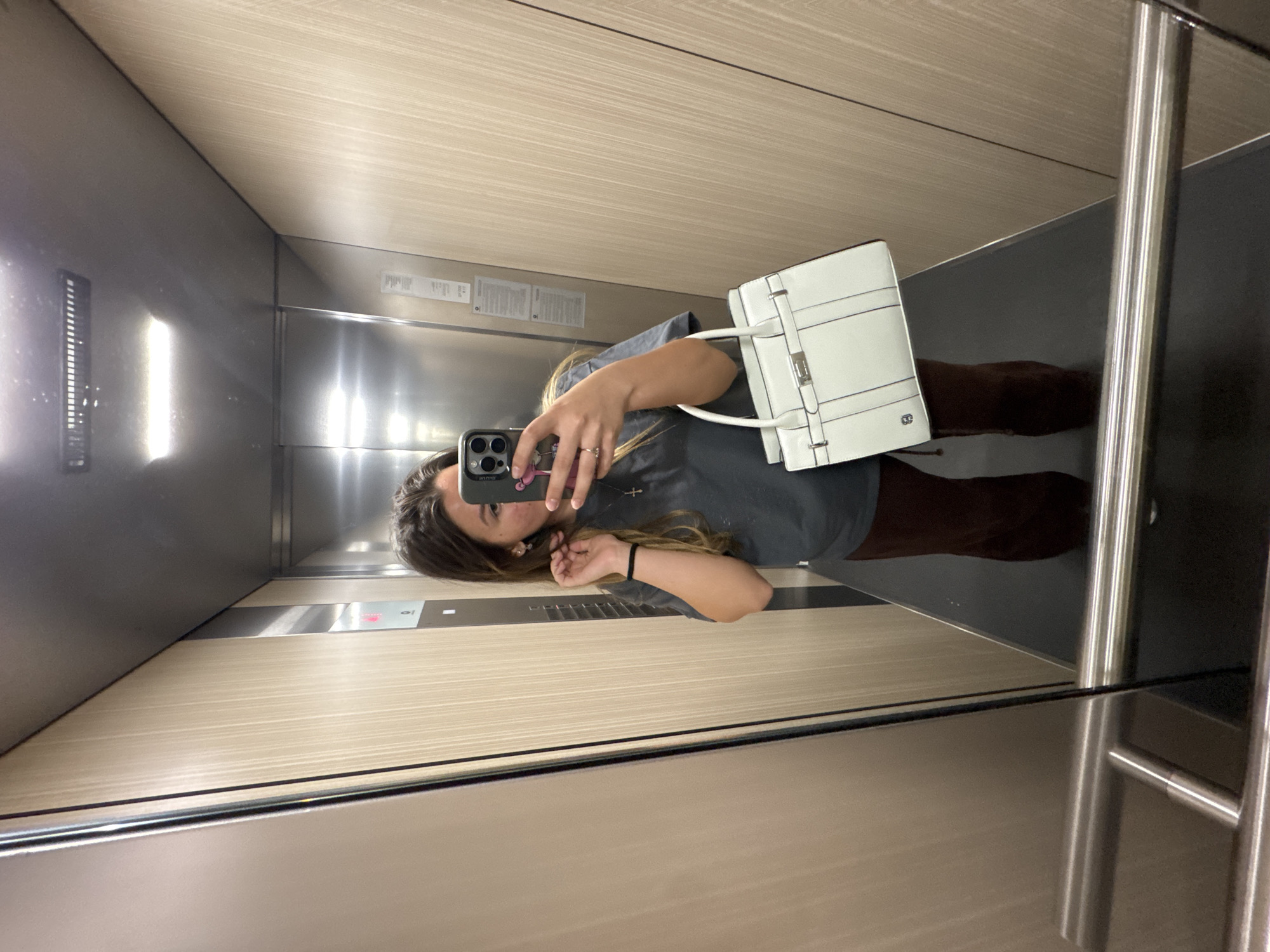 A woman stands in an elevator, holding a phone to take a selfie in the mirror. She is wearing a gray shirt and brown pants. The elevator has a wood-paneled interior and a metal door. The location is likely in the city of Sofia, Bulgaria. She is holding a white purse.