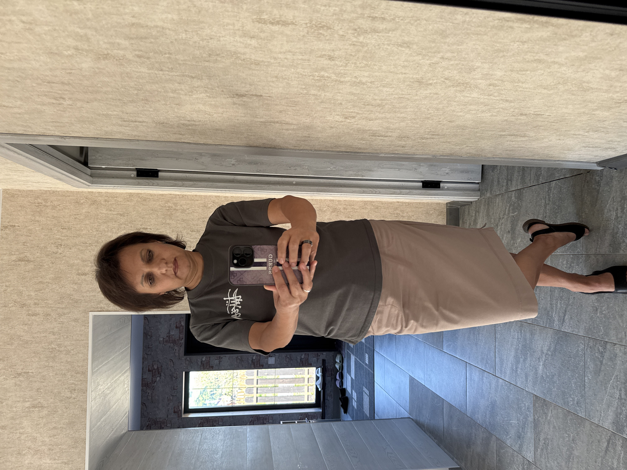 A woman stands in a hallway and takes a mirror selfie. She is wearing a grey t-shirt, a light brown skirt, and black slip-on shoes. The hallway has light grey tiled floors and beige textured walls. To her left, a doorway reveals a room with brick walls and a window overlooking a wooden fence. To her right, another doorway is partially visible. The woman is holding a smartphone in her right hand and is wearing rings on her left ring finger and right middle finger.