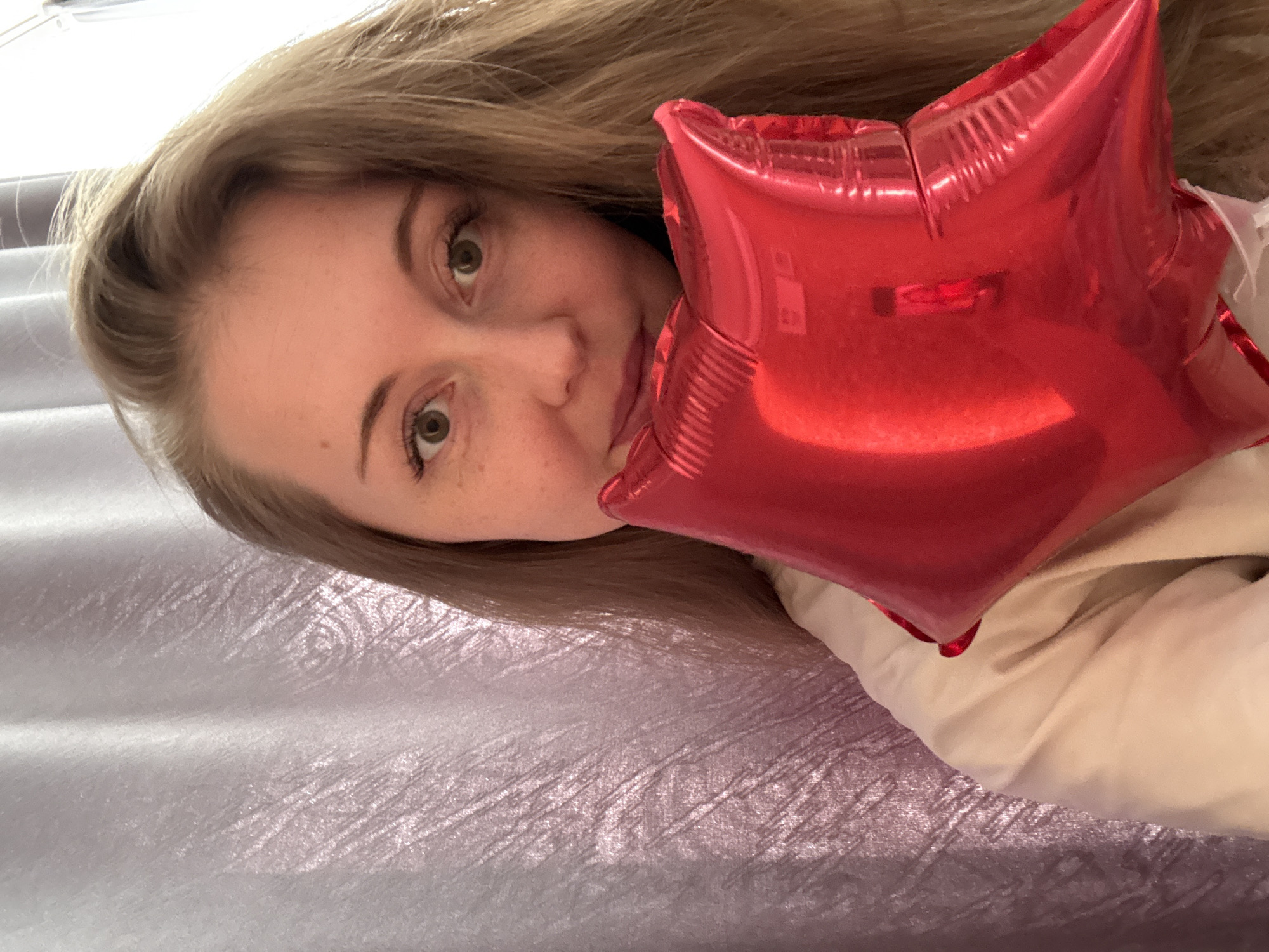A woman with light brown hair is holding a red star-shaped balloon in front of her face. She has fair skin, freckles, and light-colored eyes. The background appears to be a textured blanket or bedspread. The location is Unknown, Unknown.